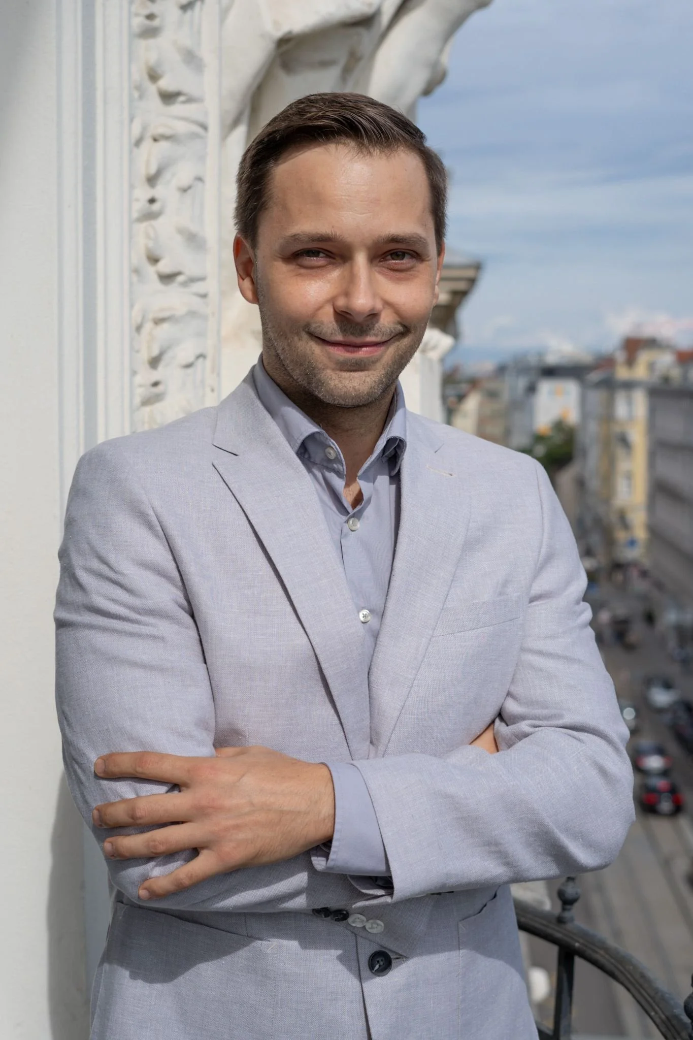 Ein junger Mann in einem hellgrauen Anzug steht auf einem Balkon mit Blick auf eine Stadt, lächelt leicht und hat die Arme verschränkt.