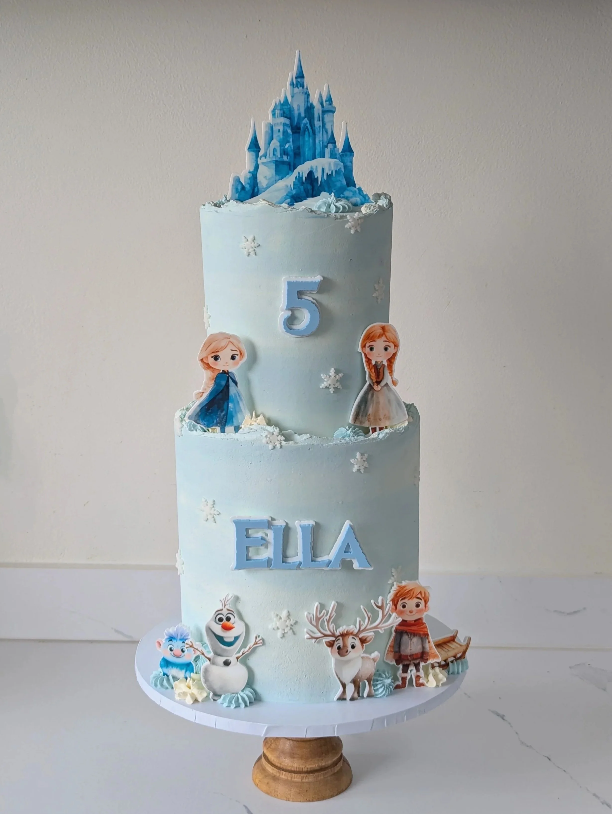 Two-tier snow-themed birthday cake decorated with Disney 'Frozen' characters—Elsa, Anna, Olaf, Sven, and others—featuring a castle topper and the name 'Ella' and number '5' on the cake.