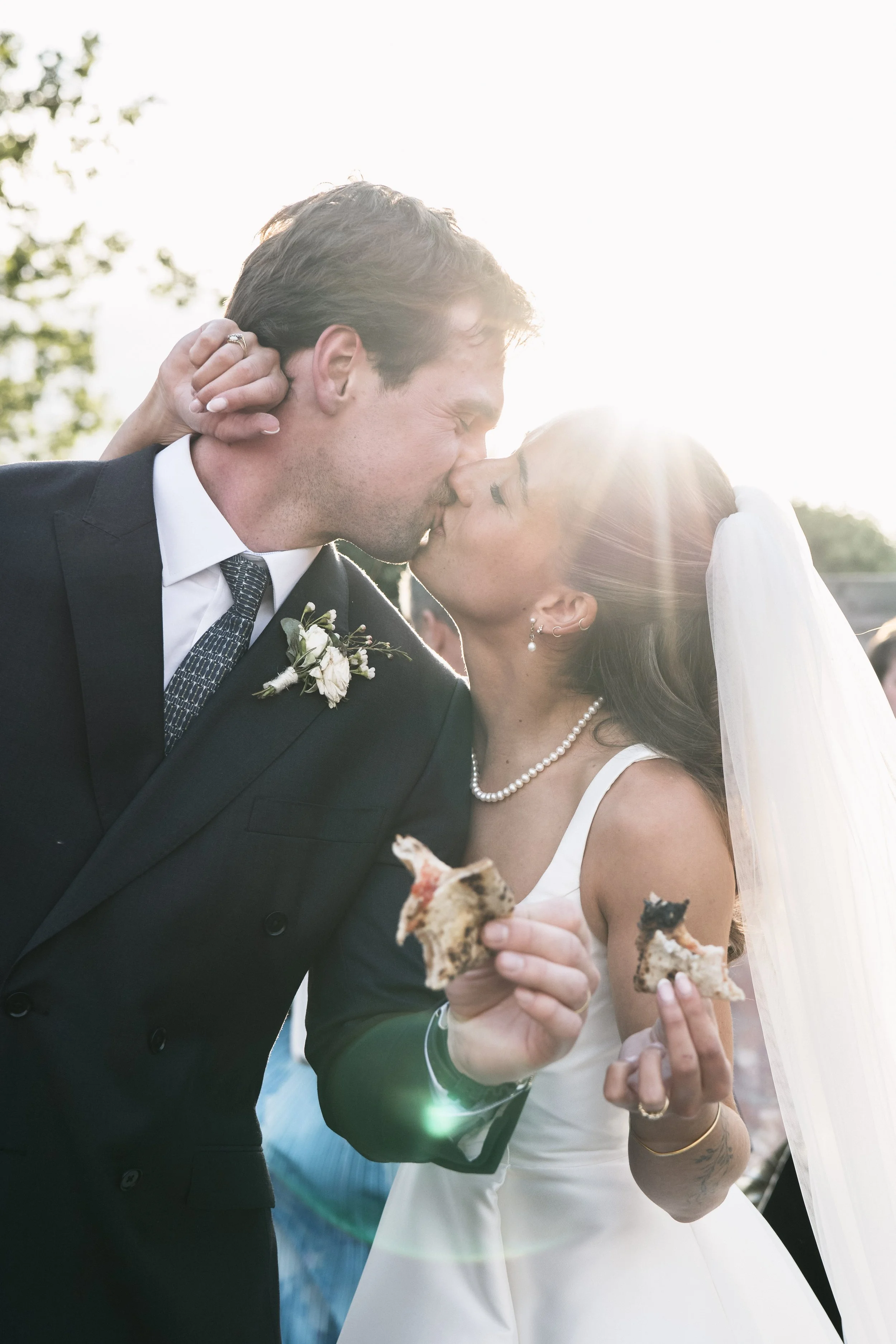 A bride and groom sharing a kiss outdoors, holding slices of pizza during their wedding celebration.