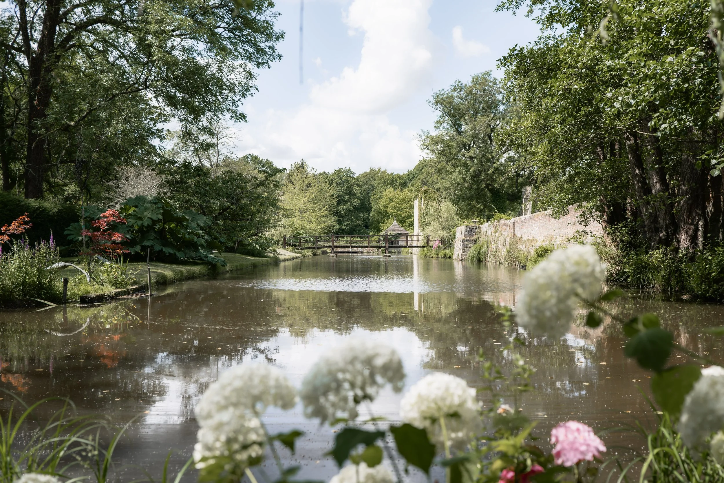 A peaceful pond surrounded by lush greenery, flowers, and trees, with a wooden bridge and gazebo in the background under a partly cloudy sky.