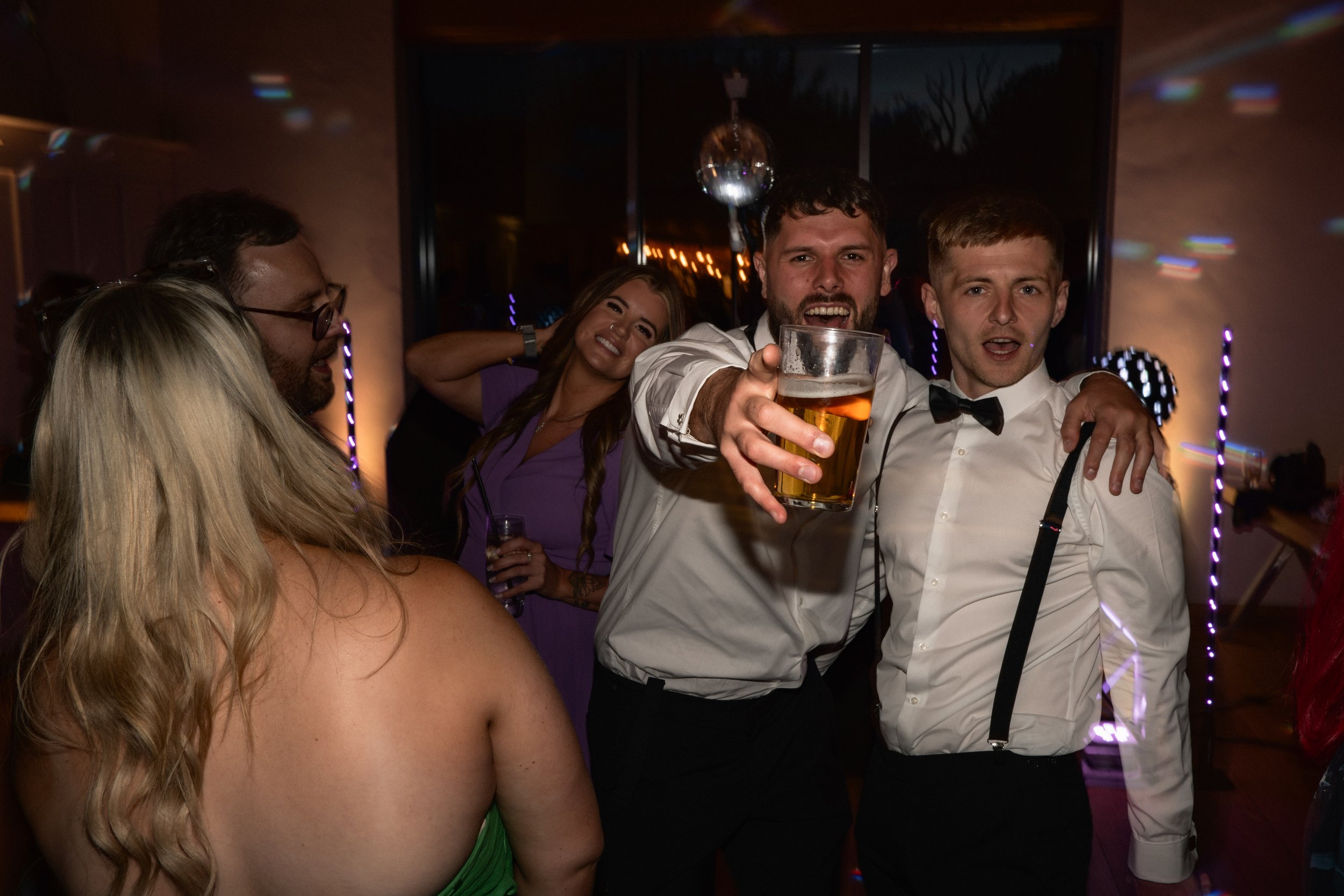 Group of friends celebrating at a party, one man is holding up a beer glass and smiling at the camera, two men are dressed in formal attire with suspenders and bow ties, women are dressed in party dresses, colorful lights and a disco ball in the back
