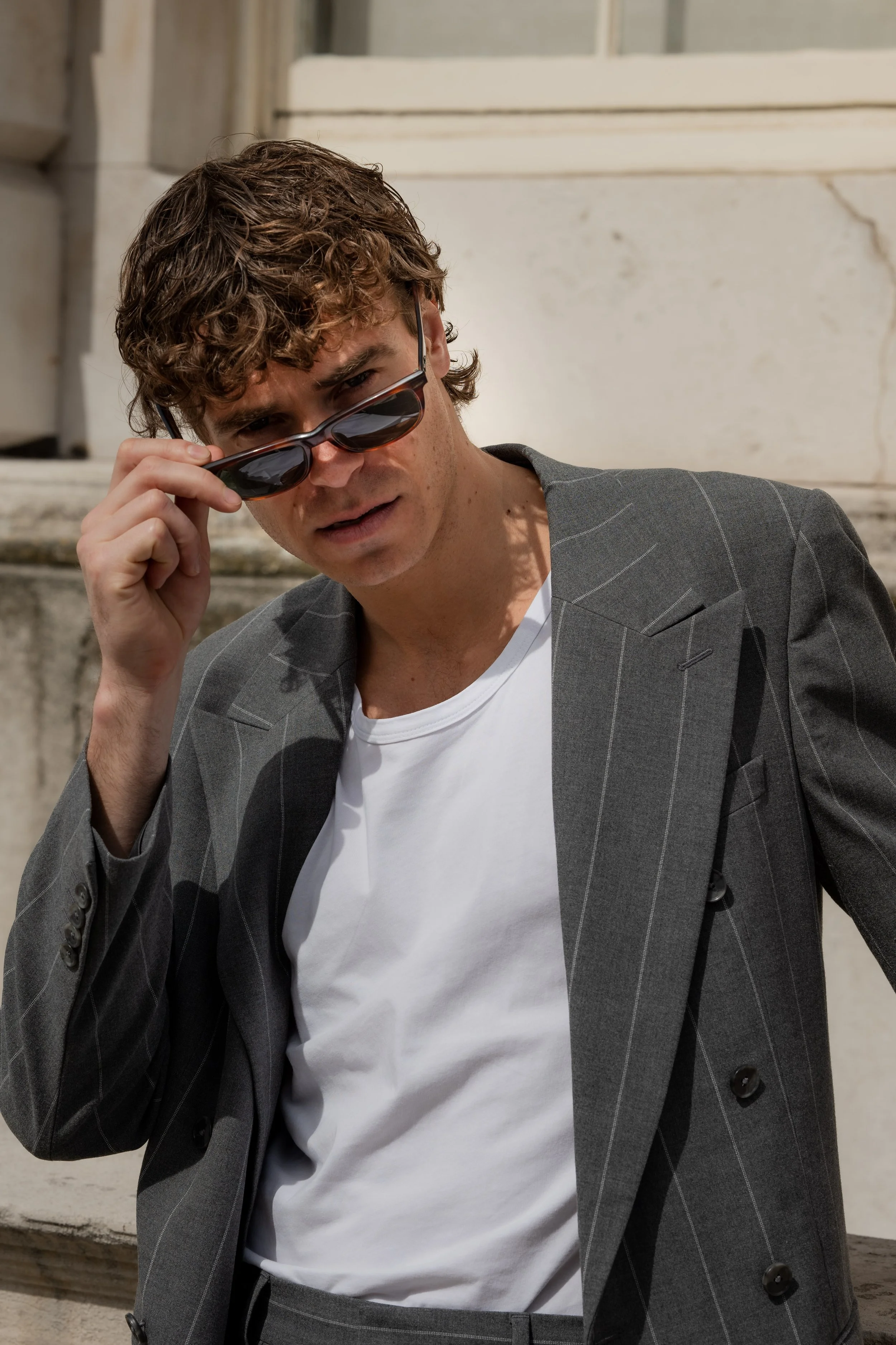 A man with curly hair wearing sunglasses, a white t-shirt, and a gray pinstripe blazer, looking over his sunglasses in front of a stone building.