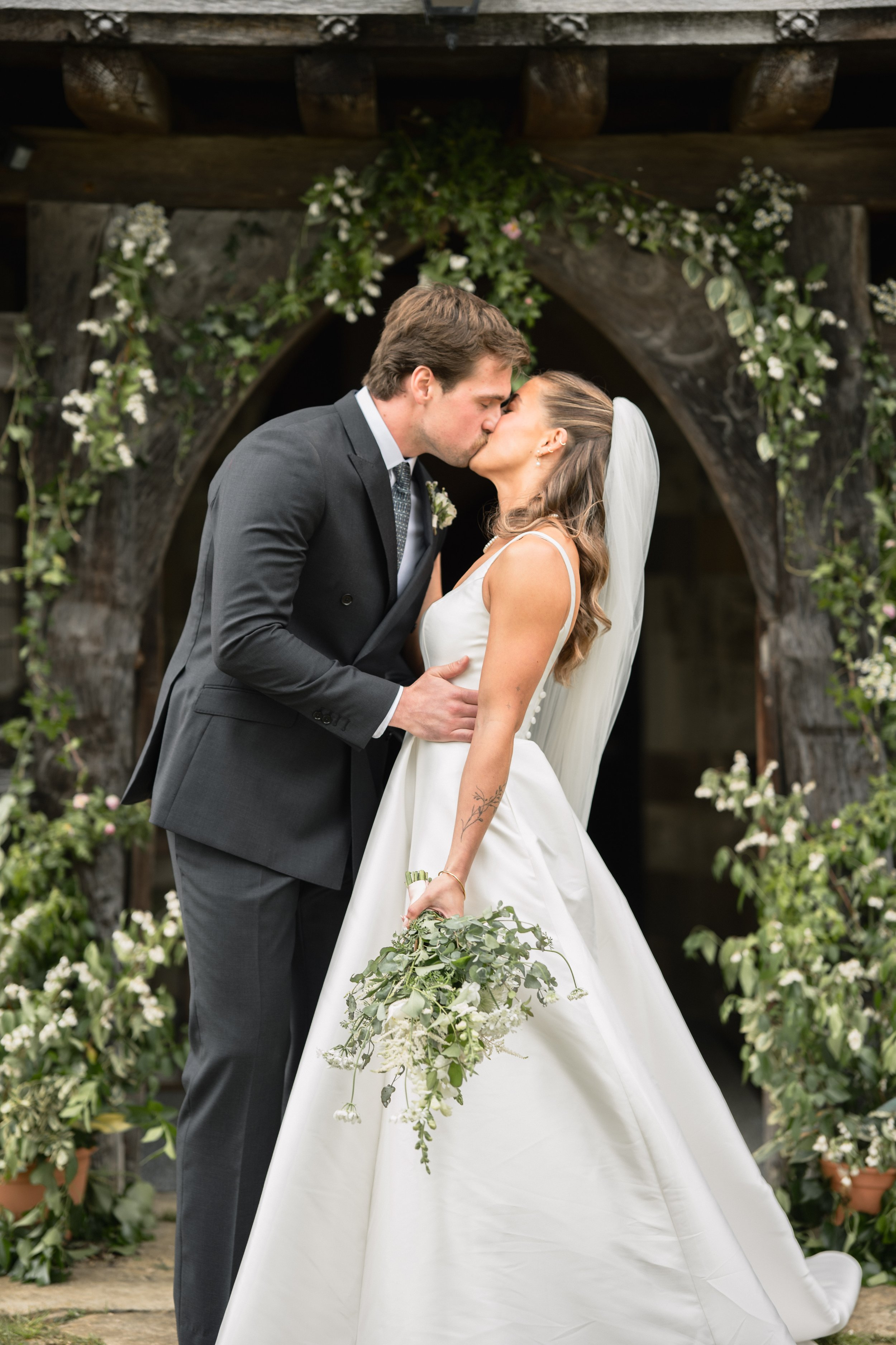 A bride and groom share a kiss during their wedding ceremony, standing under a rustic wooden arch decorated with white flowers and greenery.