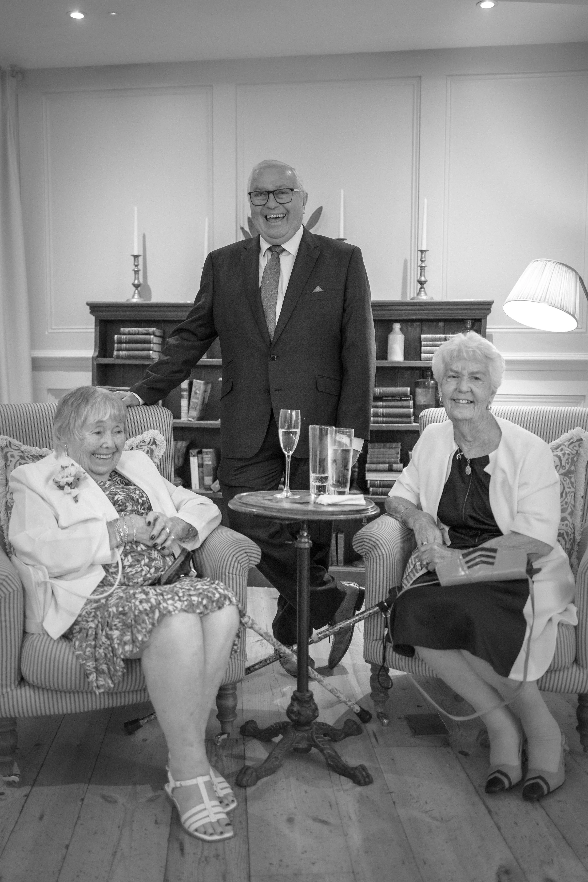 A man in a suit and glasses standing between two elderly women seated on striped armchairs in a living room, with a small round table holding glasses of drinks, and a bookshelf behind them.