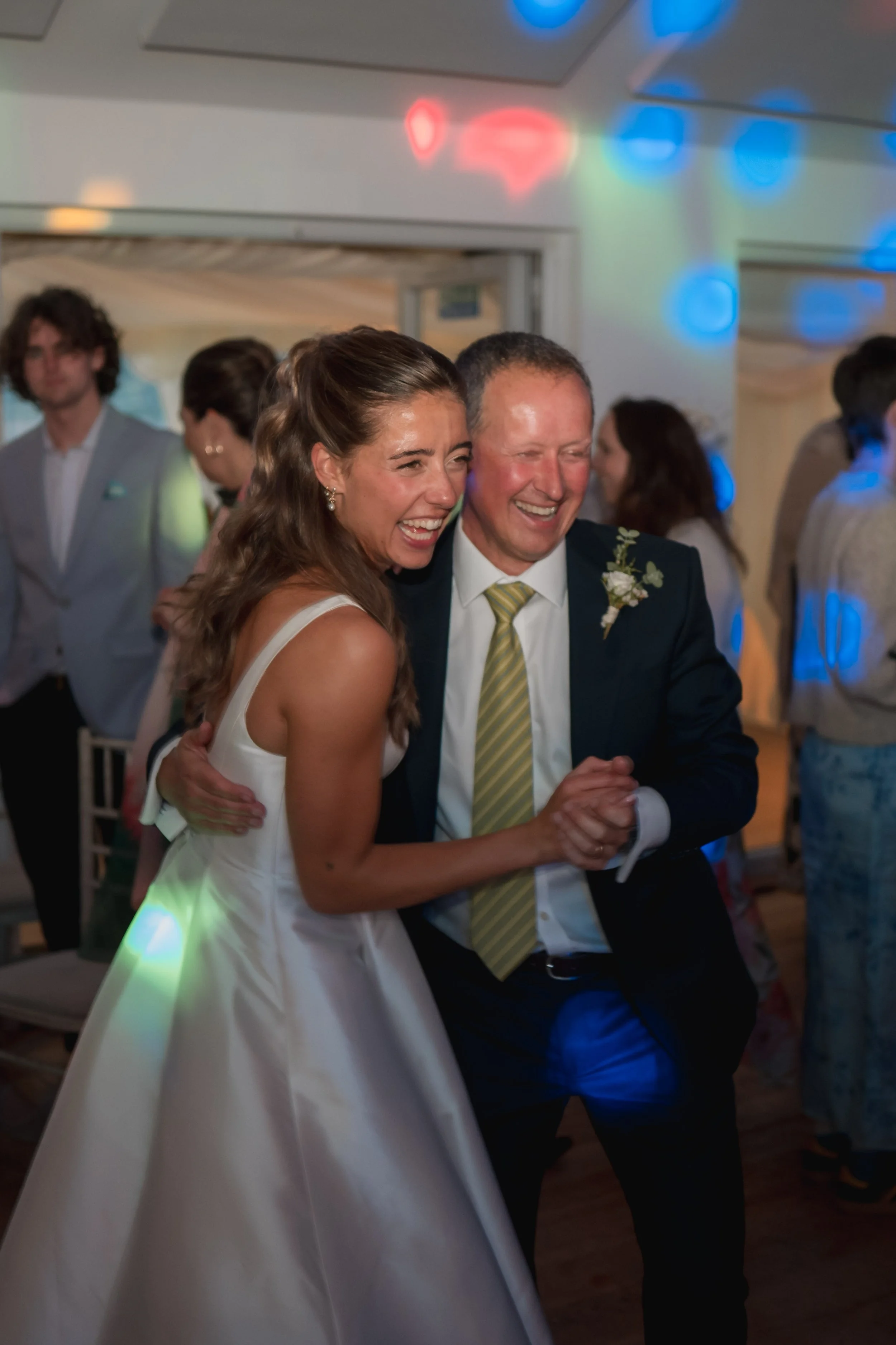 A woman in a white dress and a man in a suit are dancing together and laughing at a wedding reception, with other guests and colorful party lights in the background.