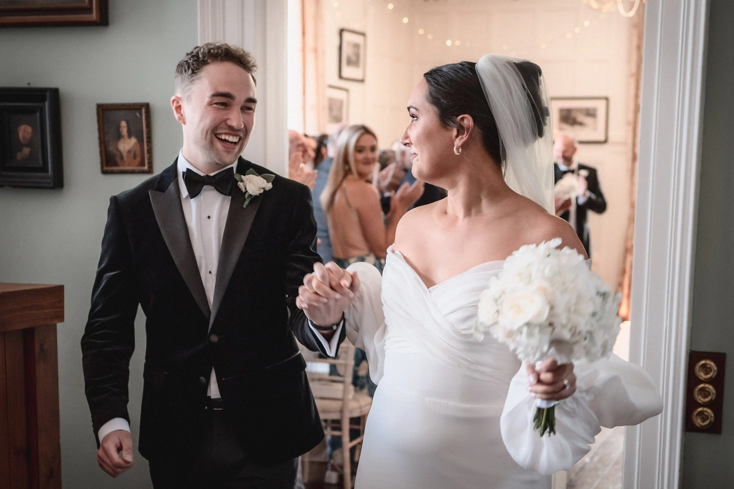 A newlywed couple celebrating their wedding, holding hands and smiling at each other in an indoor setting with guests in the background.