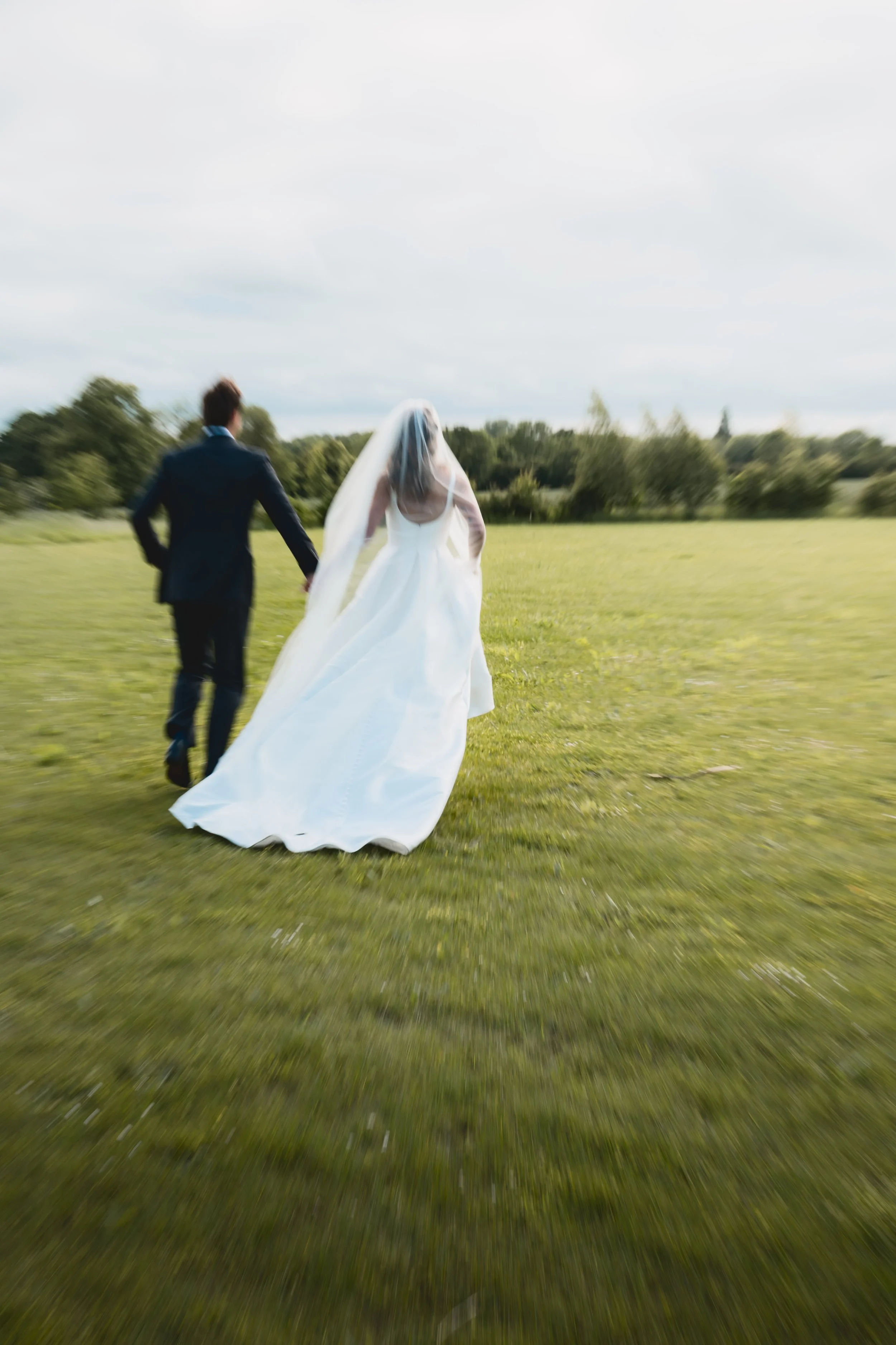 A bride and groom walking hand in hand across a grassy field, with trees in the background and the sky above.