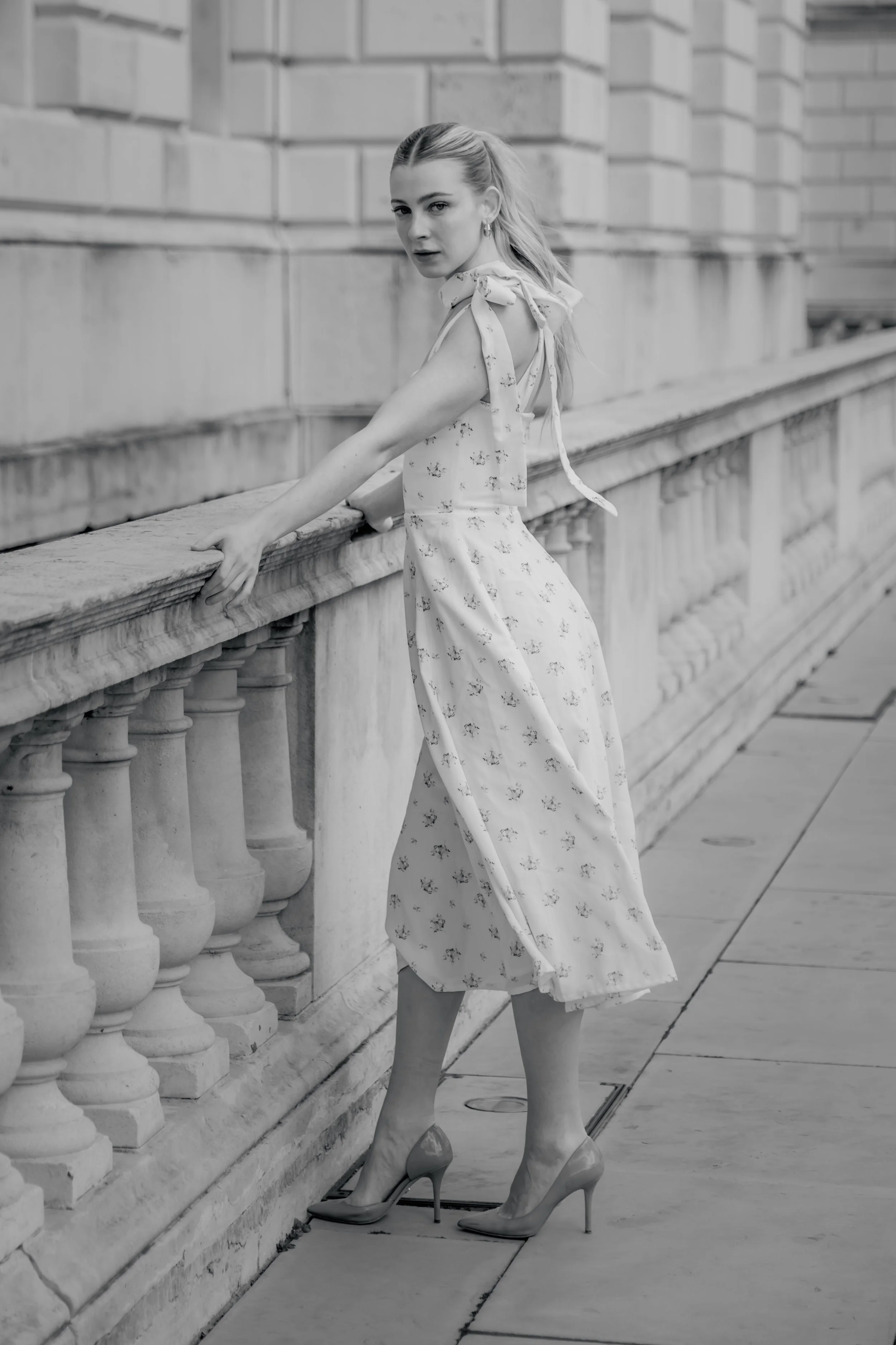 A woman in a floral dress and high heels standing outdoors near a stone railing, looking over her shoulder at the camera, in black and white.