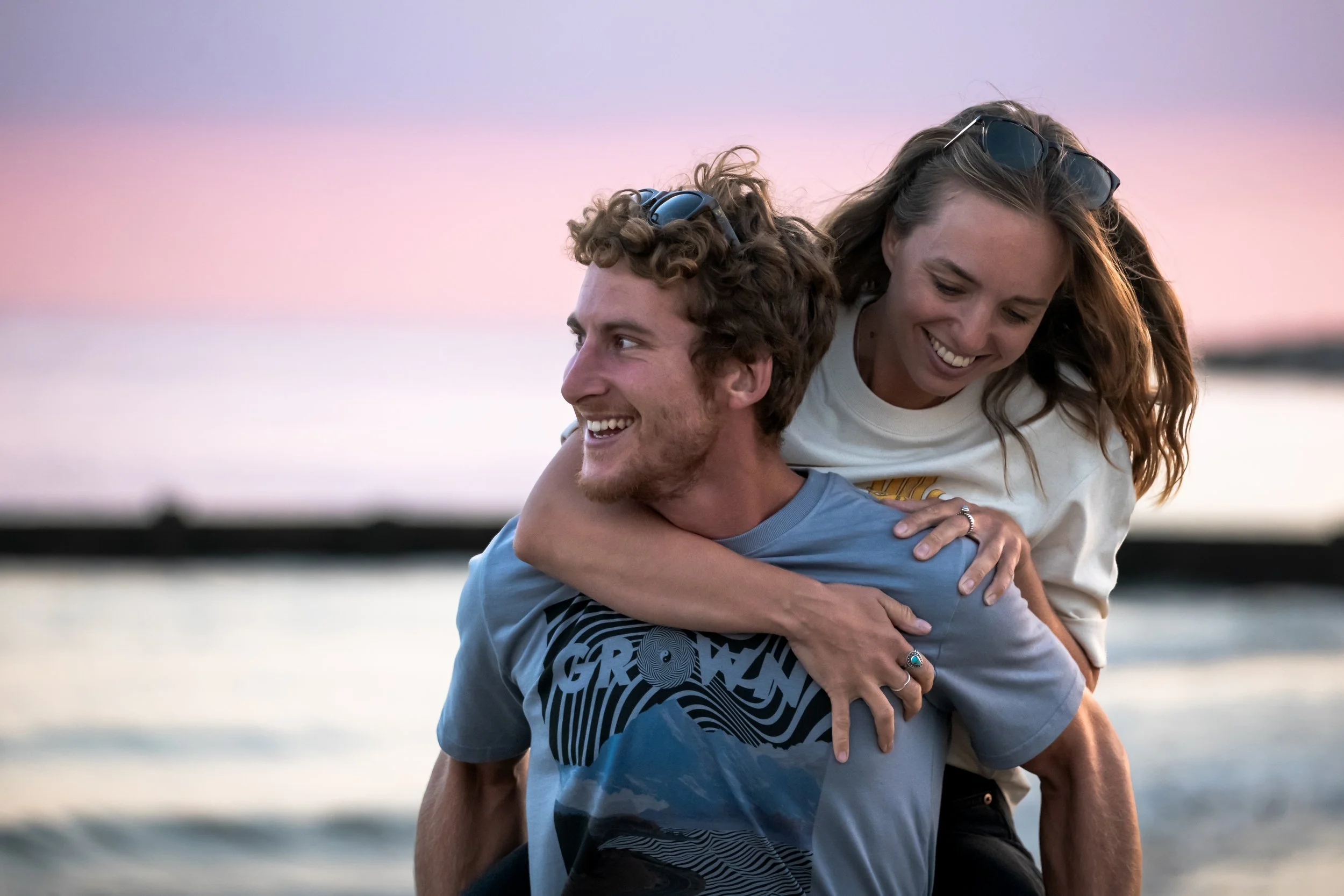 A young man giving a piggyback ride to a young woman on a beach at sunset, both smiling and enjoying themselves.