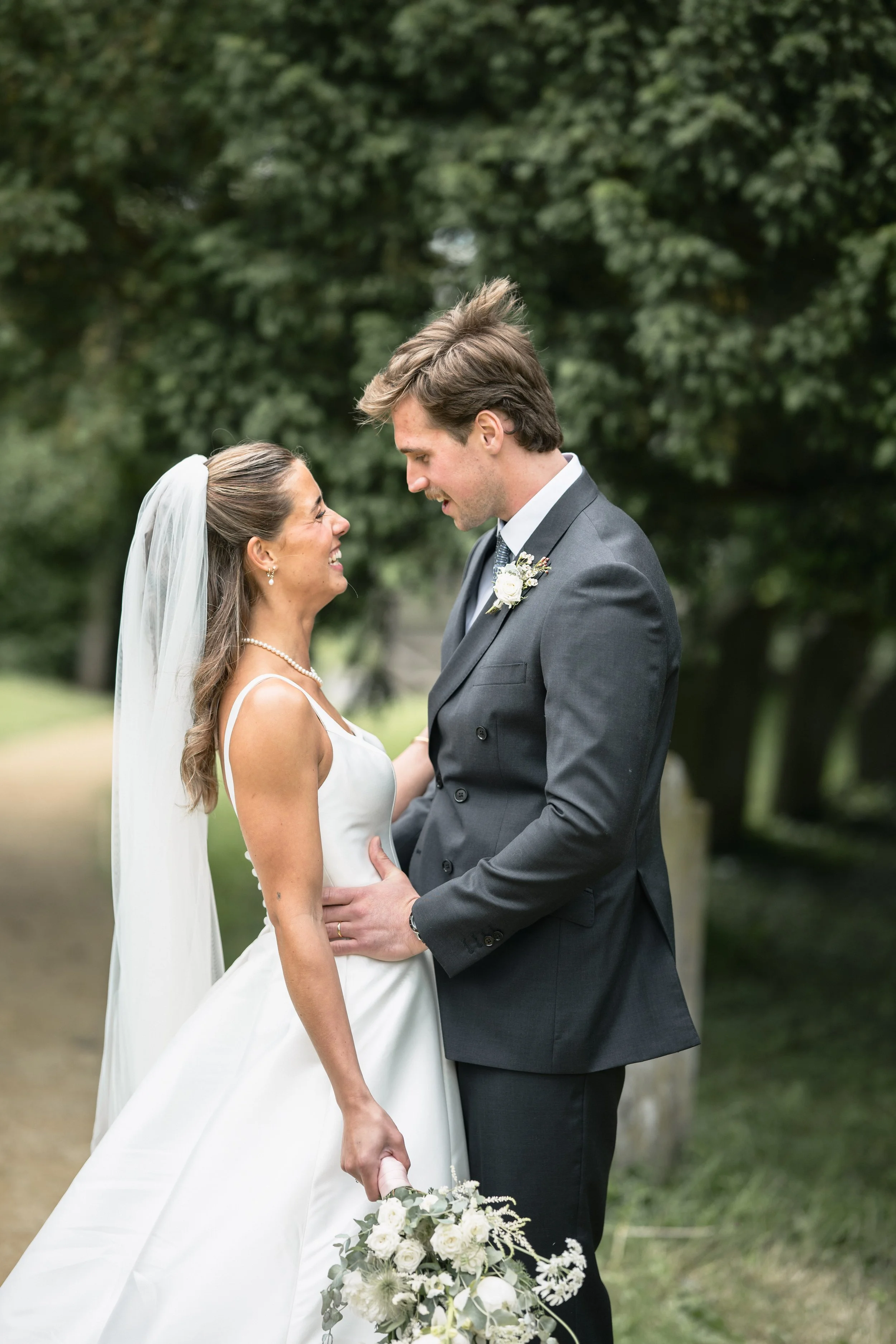 A bride and groom standing outdoors, smiling and looking into each other's eyes, with trees in the background. The bride is holding a bouquet of white flowers and wearing a white wedding dress with a veil. The groom is dressed in a dark suit with a b