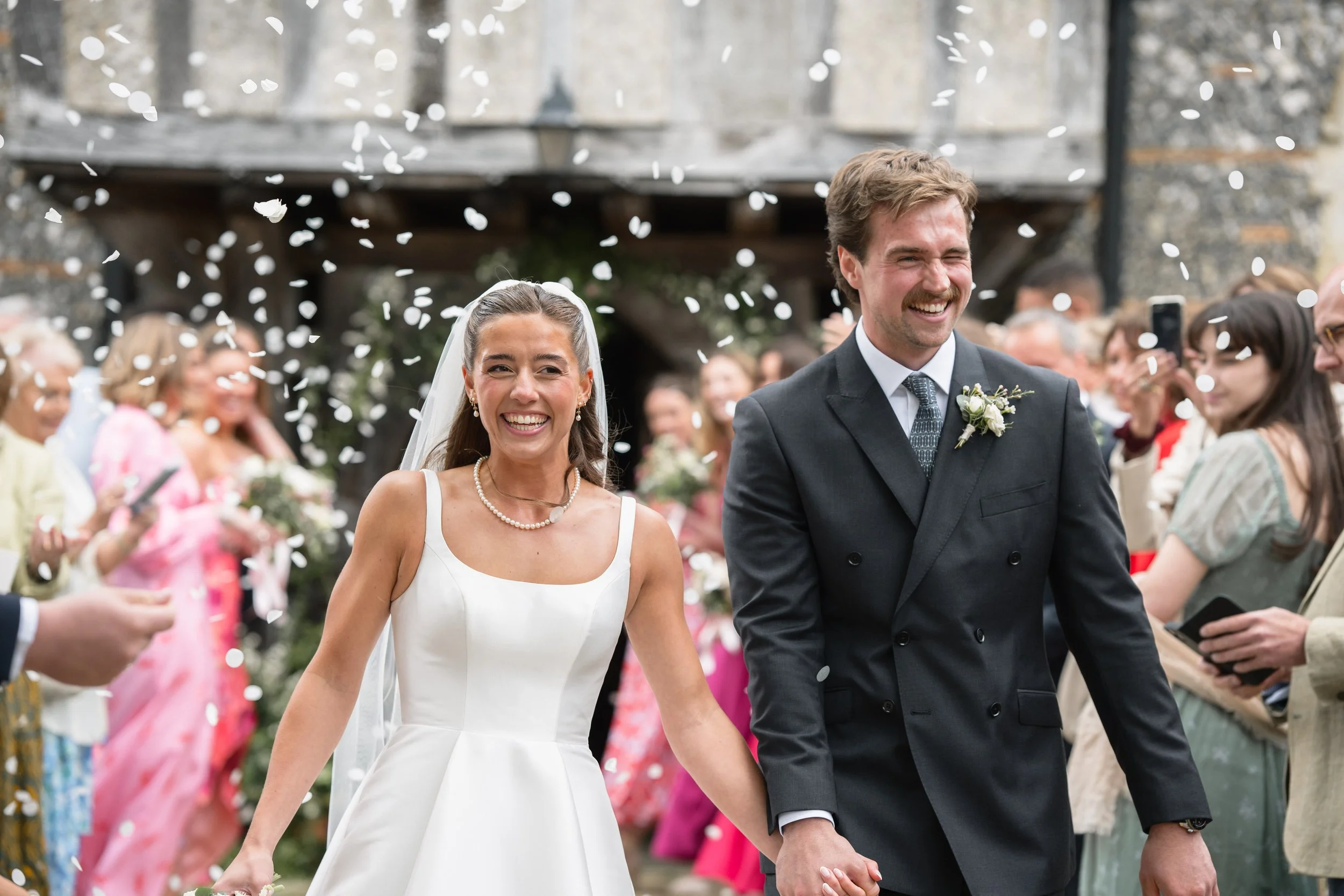 Happy newlywed couple holding hands and walking outdoors after wedding ceremony, surrounded by friends and family, with confetti falling around them.