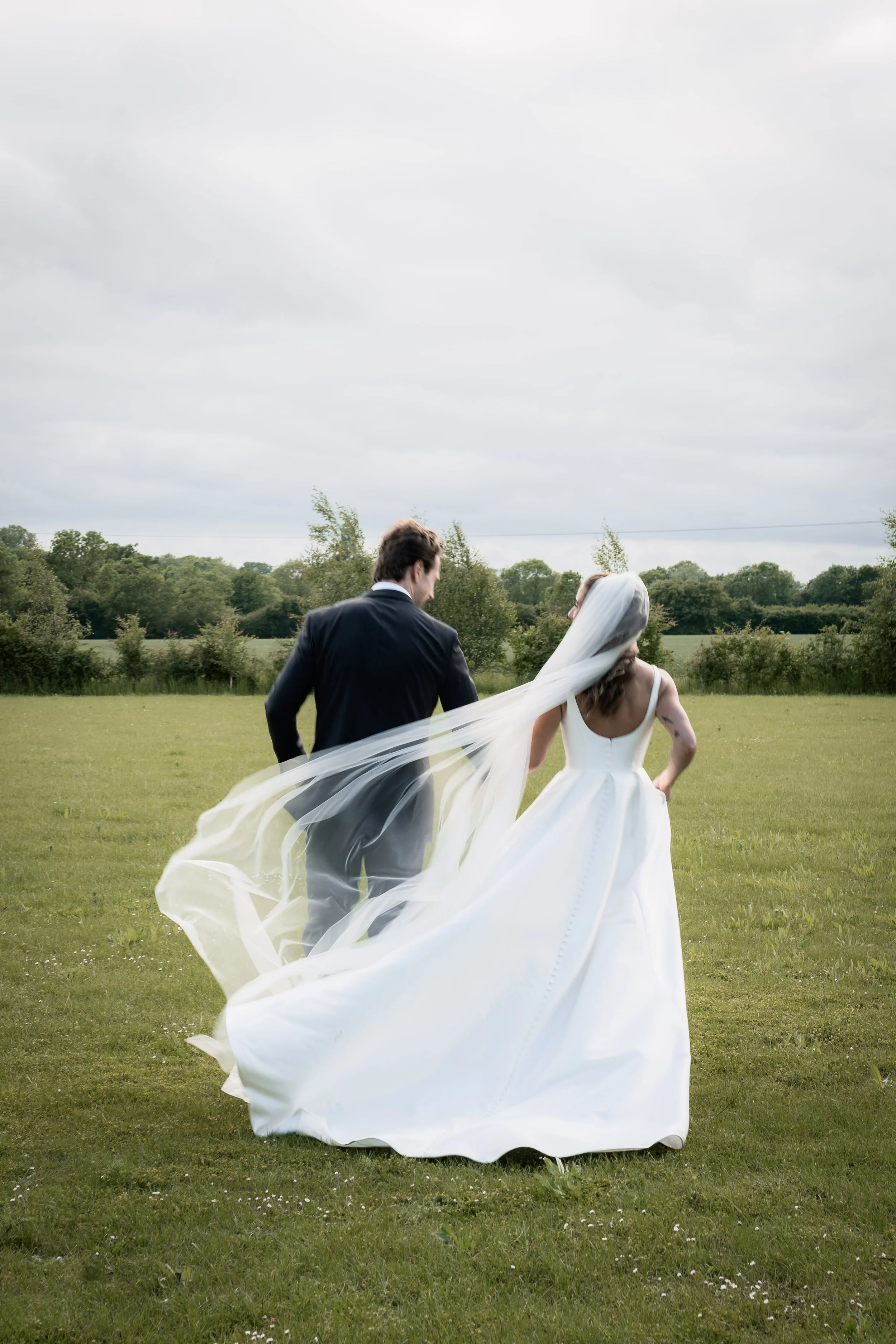 A bride and groom walking together in a grassy field, with the bride wearing a white wedding dress and veil, and the groom in a black suit, under a cloudy sky.