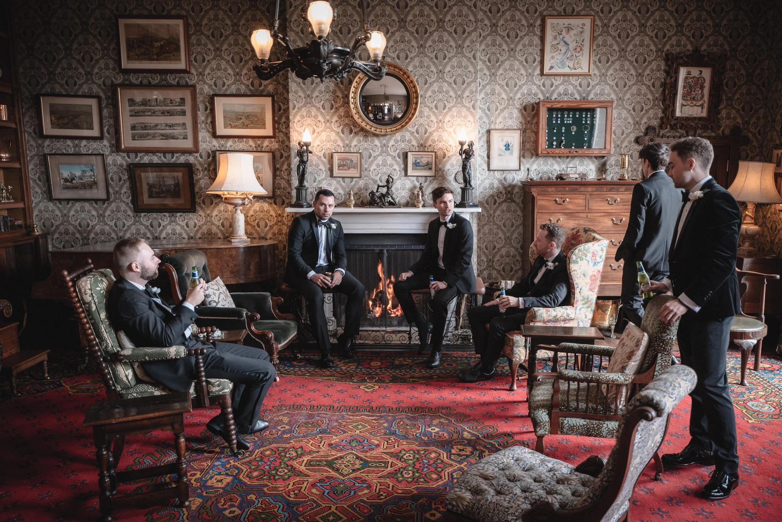 Six men in tuxedos relaxing in a vintage living room with a fireplace, ornate wallpaper, framed artwork, and antique furniture, holding drinks.