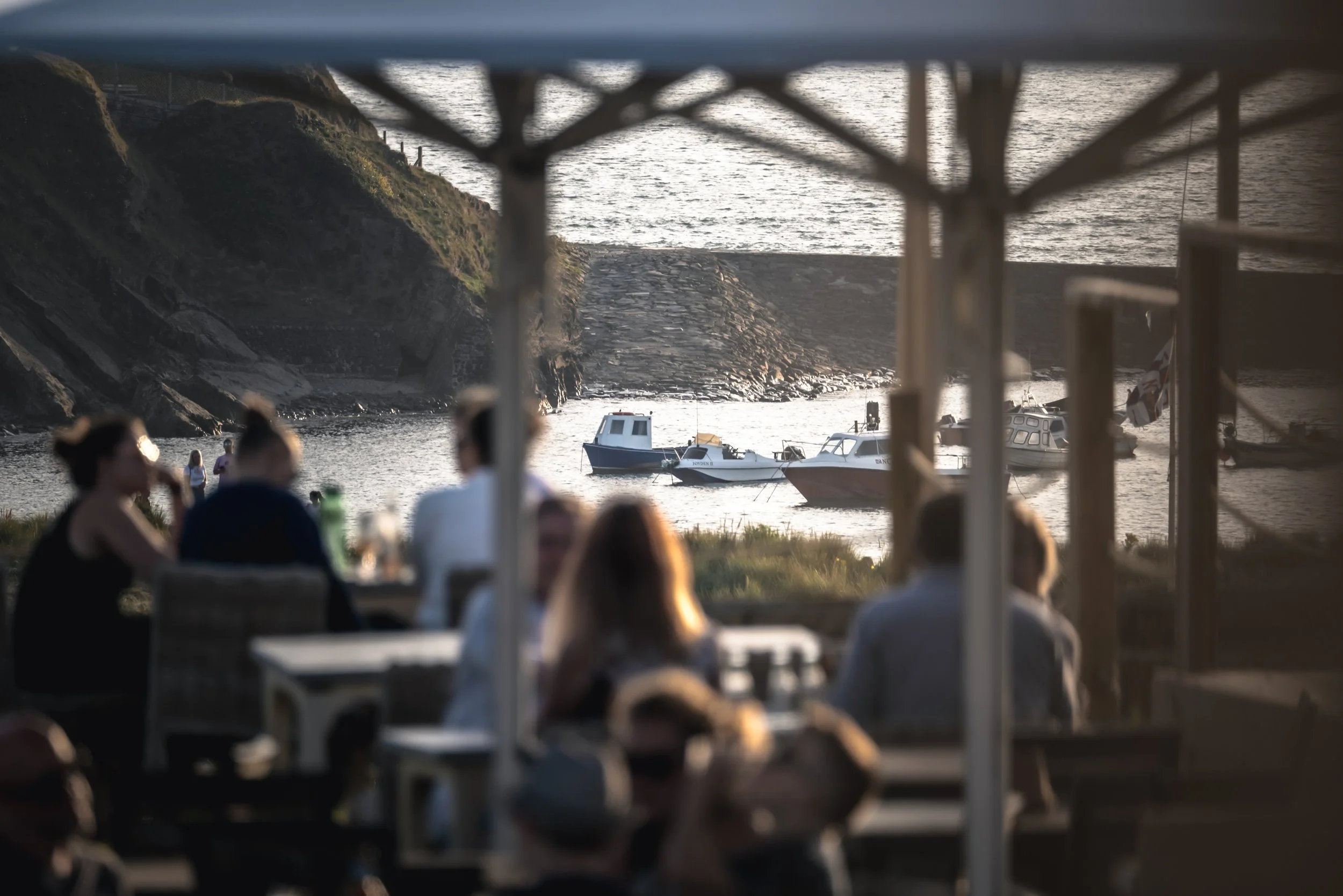People sitting at tables under a canopy near a bay with boats anchored in the water and a rocky hillside in the background during sunset.