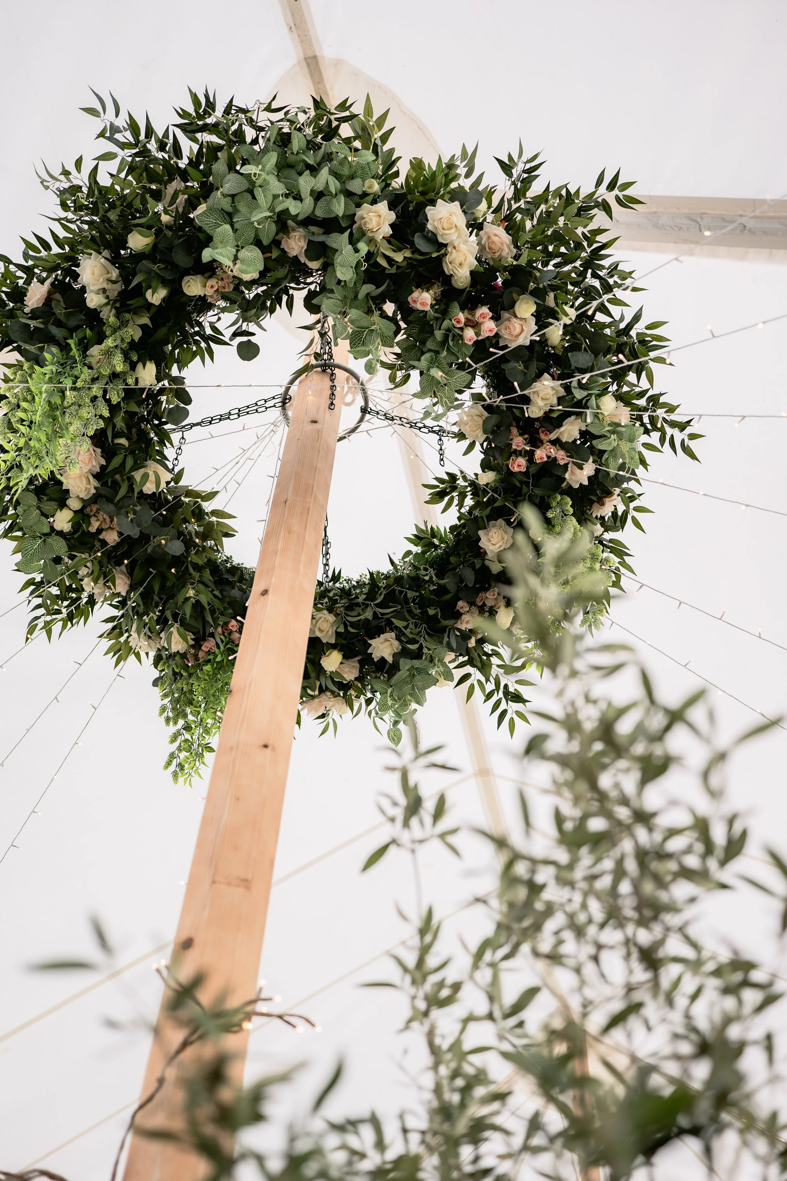 A wooden pole with a circular floral arrangement of white and pink roses and green foliage, decorated with strings of fairy lights.