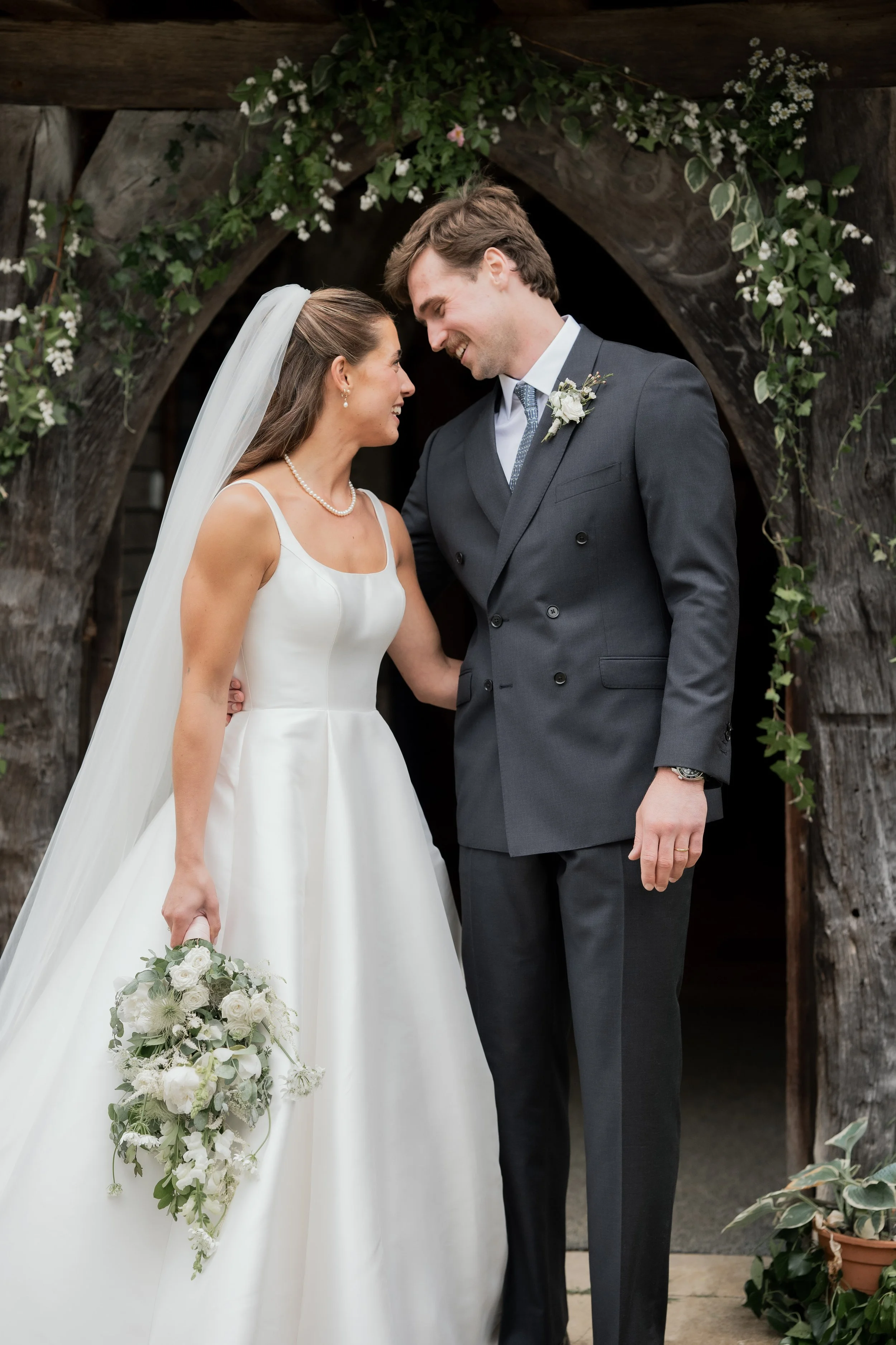 Bride and groom smiling and looking at each other, standing under a wooden arch decorated with greenery and white flowers, during their wedding ceremony.