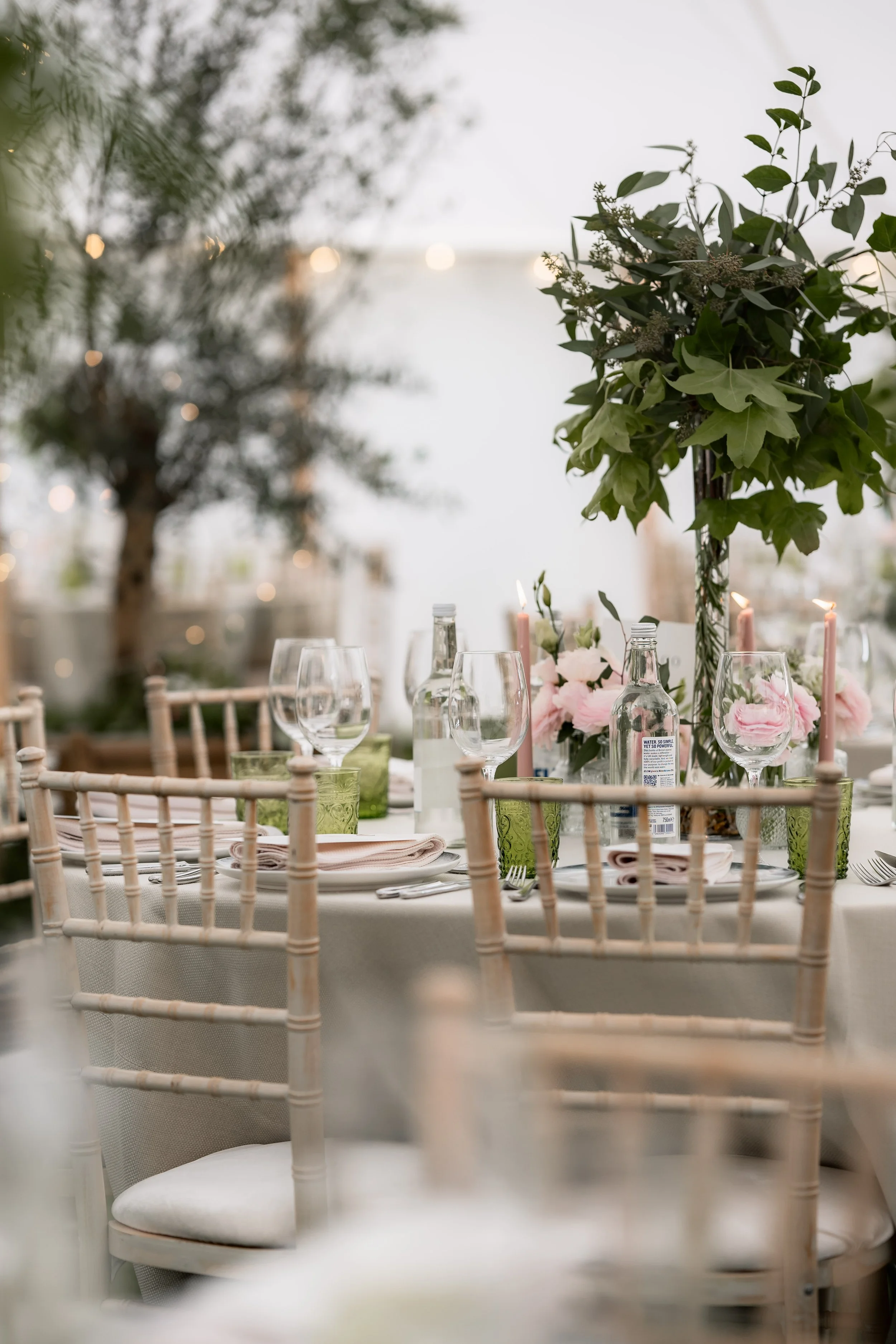 Elegant outdoor dining table set with glassware, pink flowers, candles, and greenery
