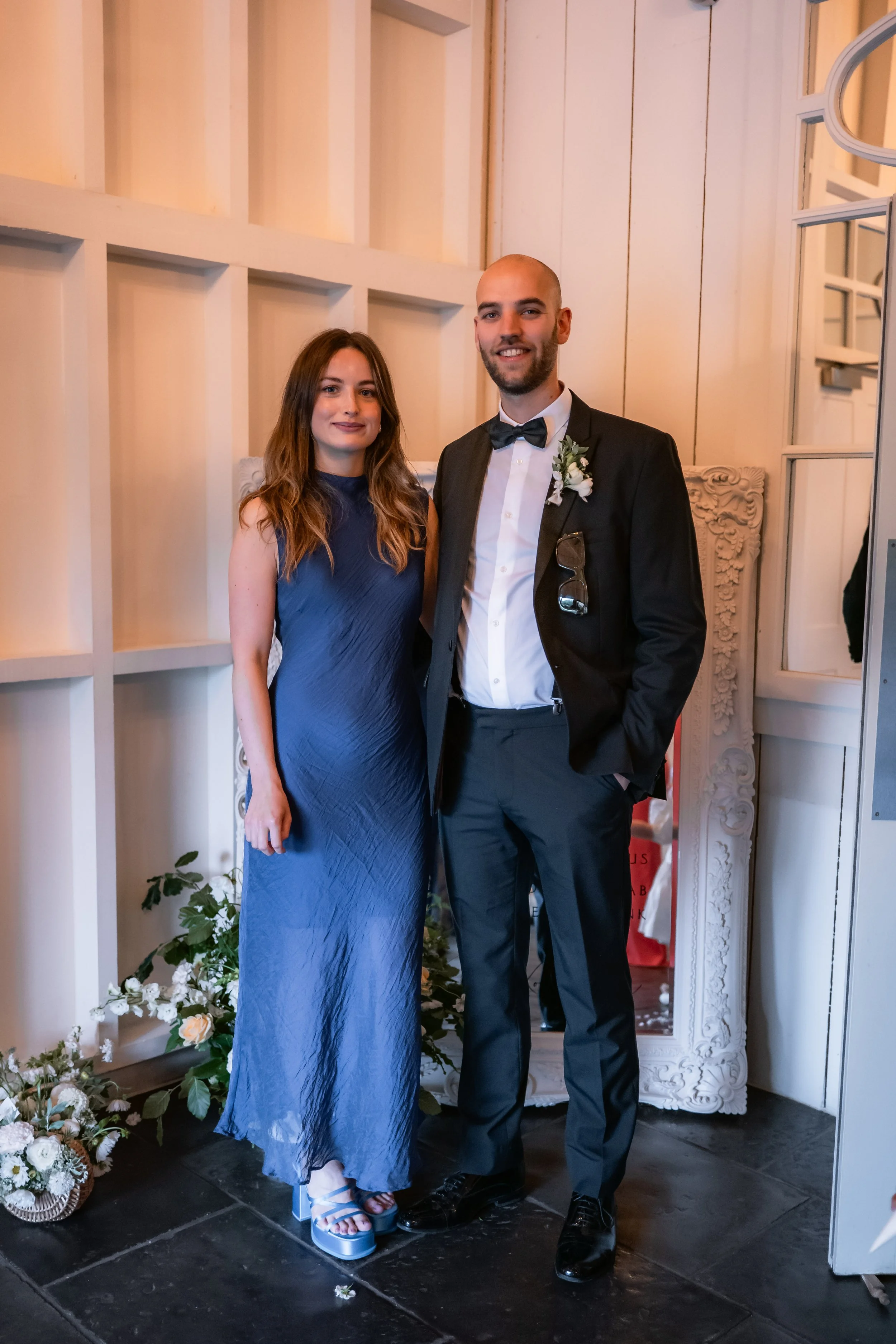 A man in a tuxedo and a woman in a blue dress standing together indoors, smiling at the camera. The man has a boutonniere and sunglasses hanging from his jacket, suggesting a formal event or celebration.