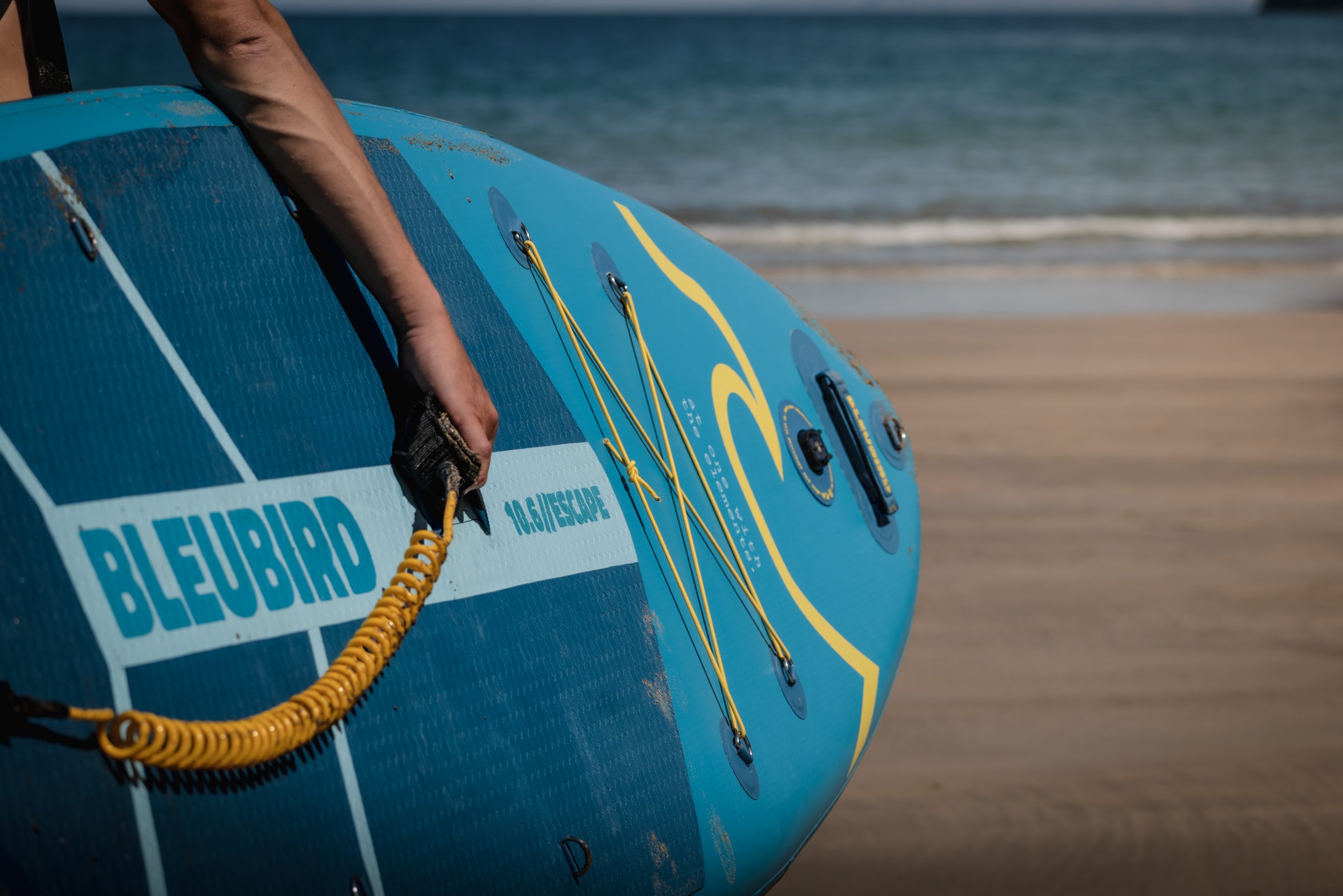 A blue and yellow inflatable paddleboard with a person's hand holding a yellow coiled leash, on the sandy beach near the ocean.