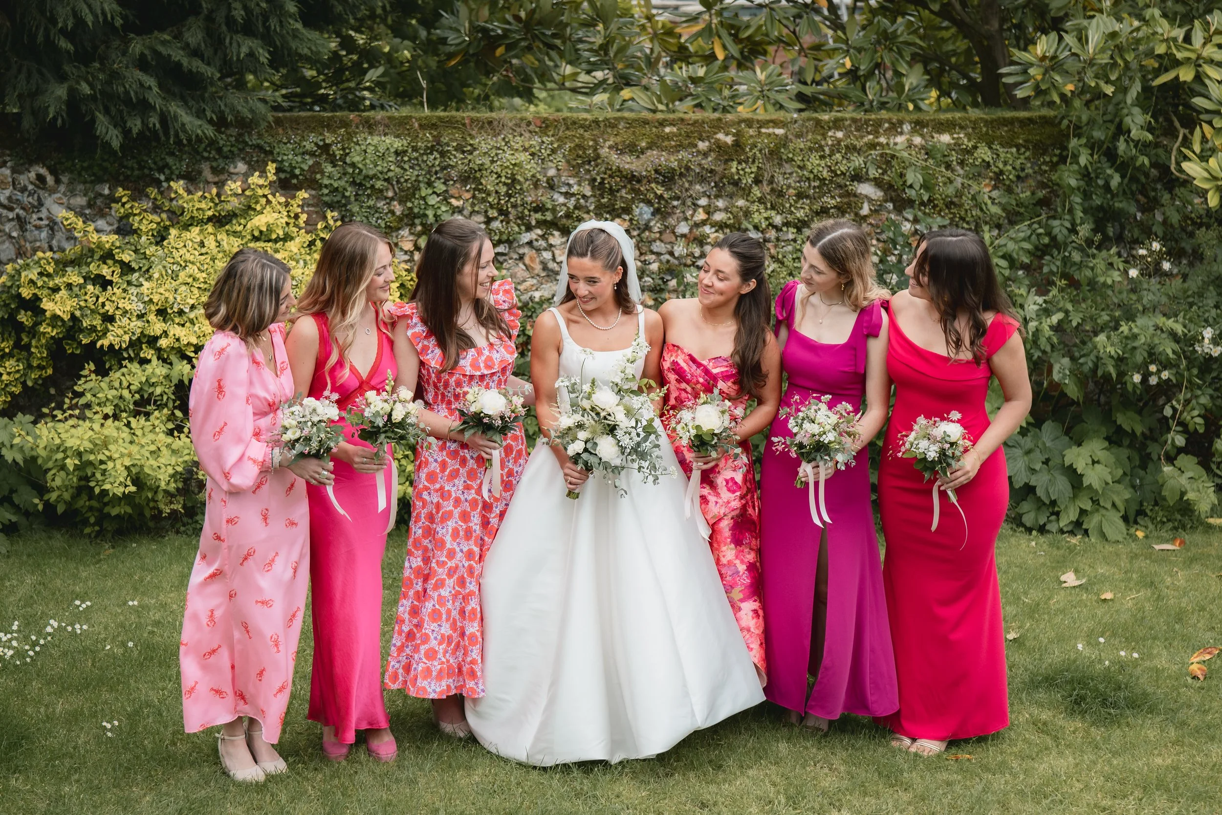 A bride in a white wedding dress standing with six women in colorful dresses holding bouquets in a garden setting.