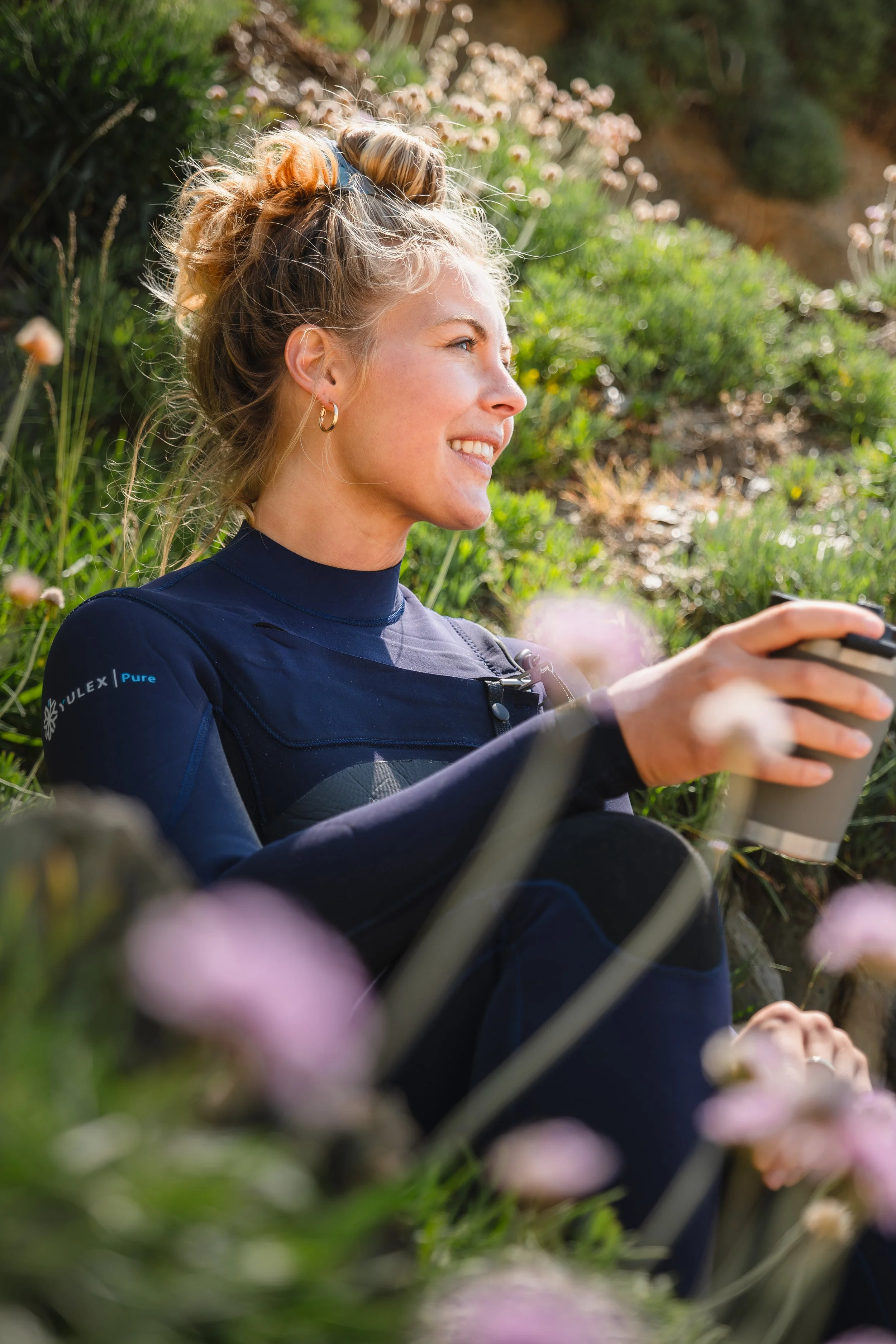 A young woman with blonde hair tied up in a bun, smiling and sitting outdoors in a lush, flowered setting, holding a travel mug.