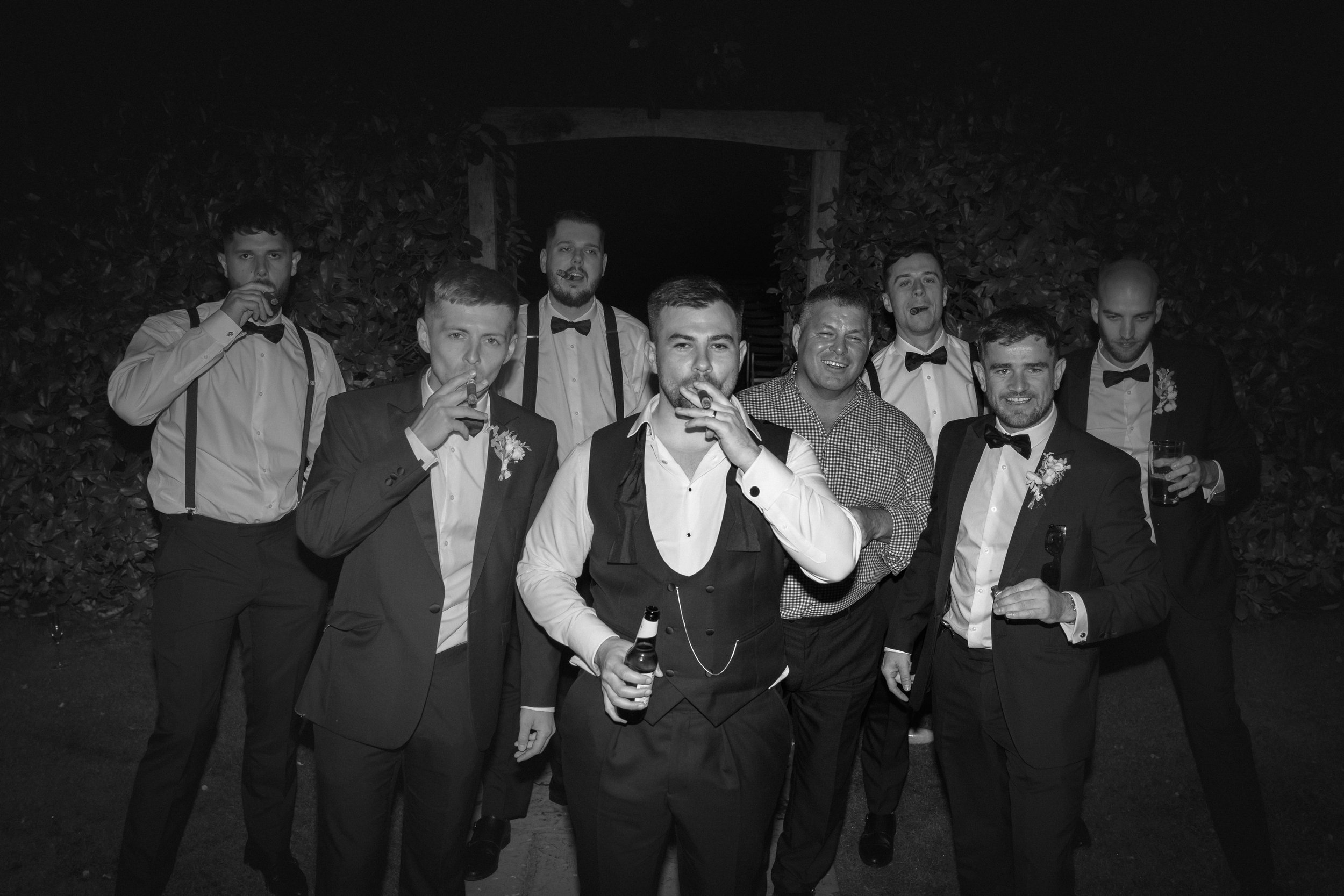 Men in formal suits and bow ties at an evening outdoor gathering, some holding drinks and cigars, smiling and posing for the photo.