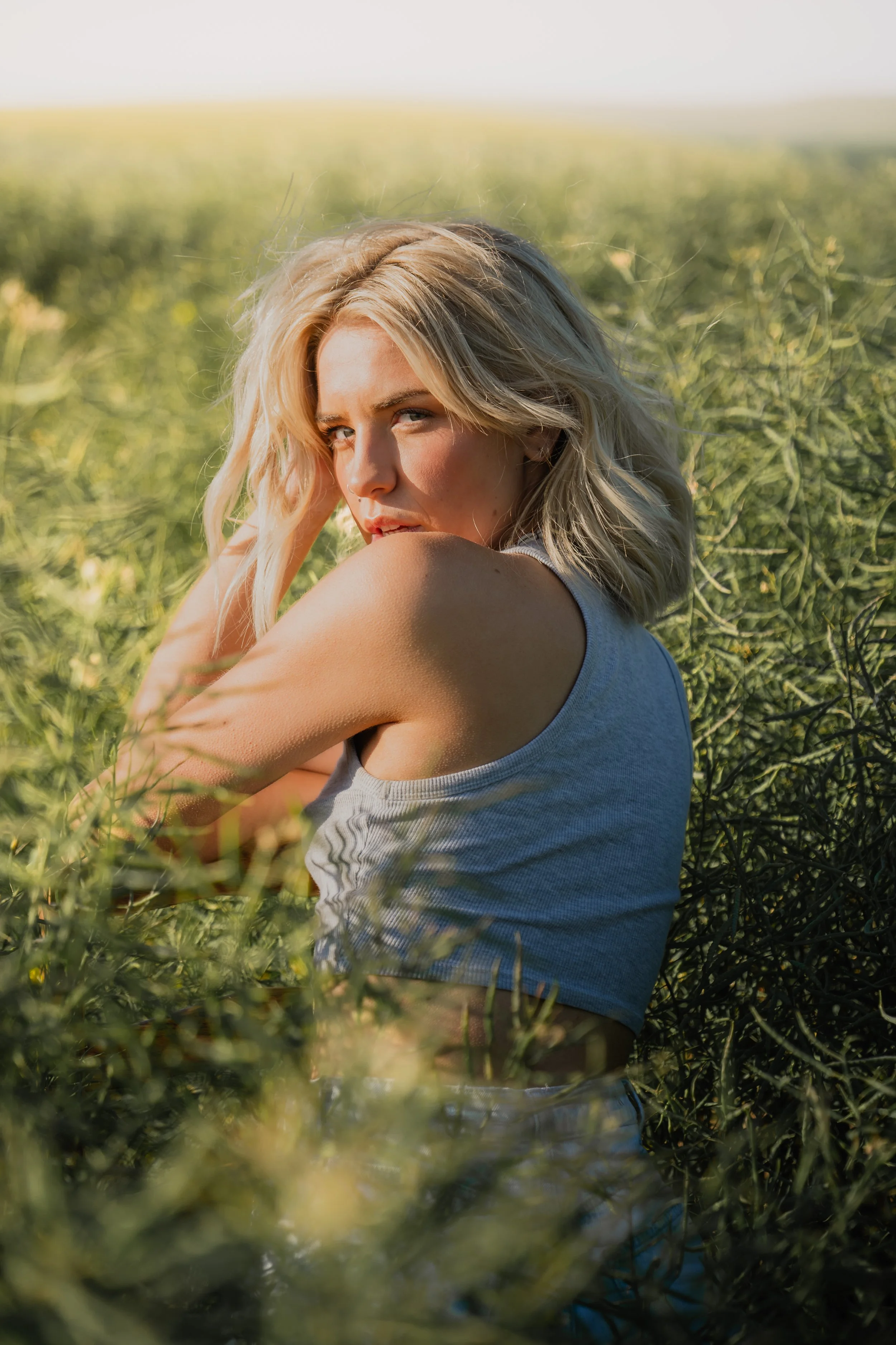 A woman with blonde hair and light skin sitting in a field of tall green grass or plants, wearing a sleeveless gray top, with the sun illuminating her face and hair.
