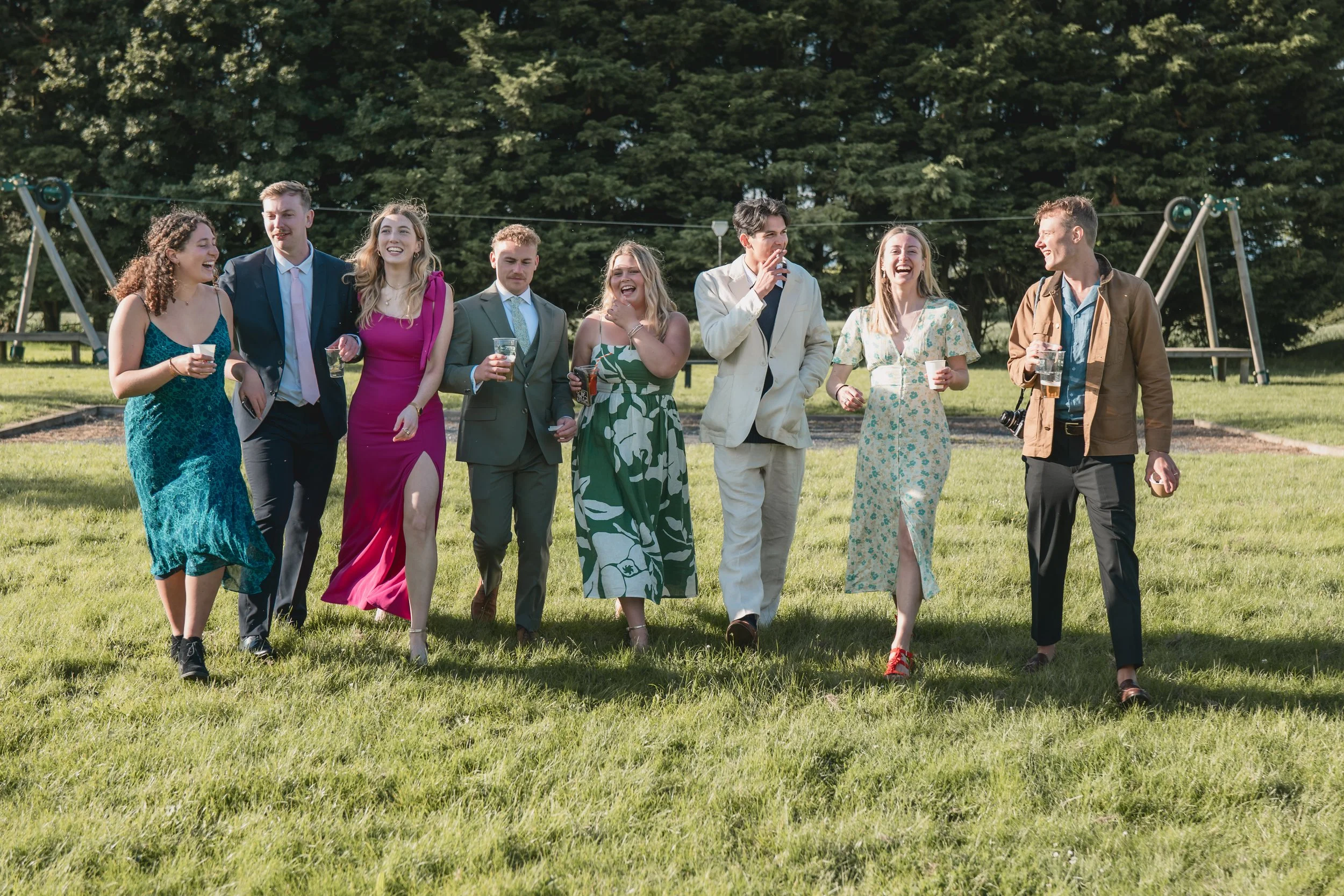 Group of nine friends walking on a grassy field, enjoying drinks, with children’s swings in the background, during daytime.
