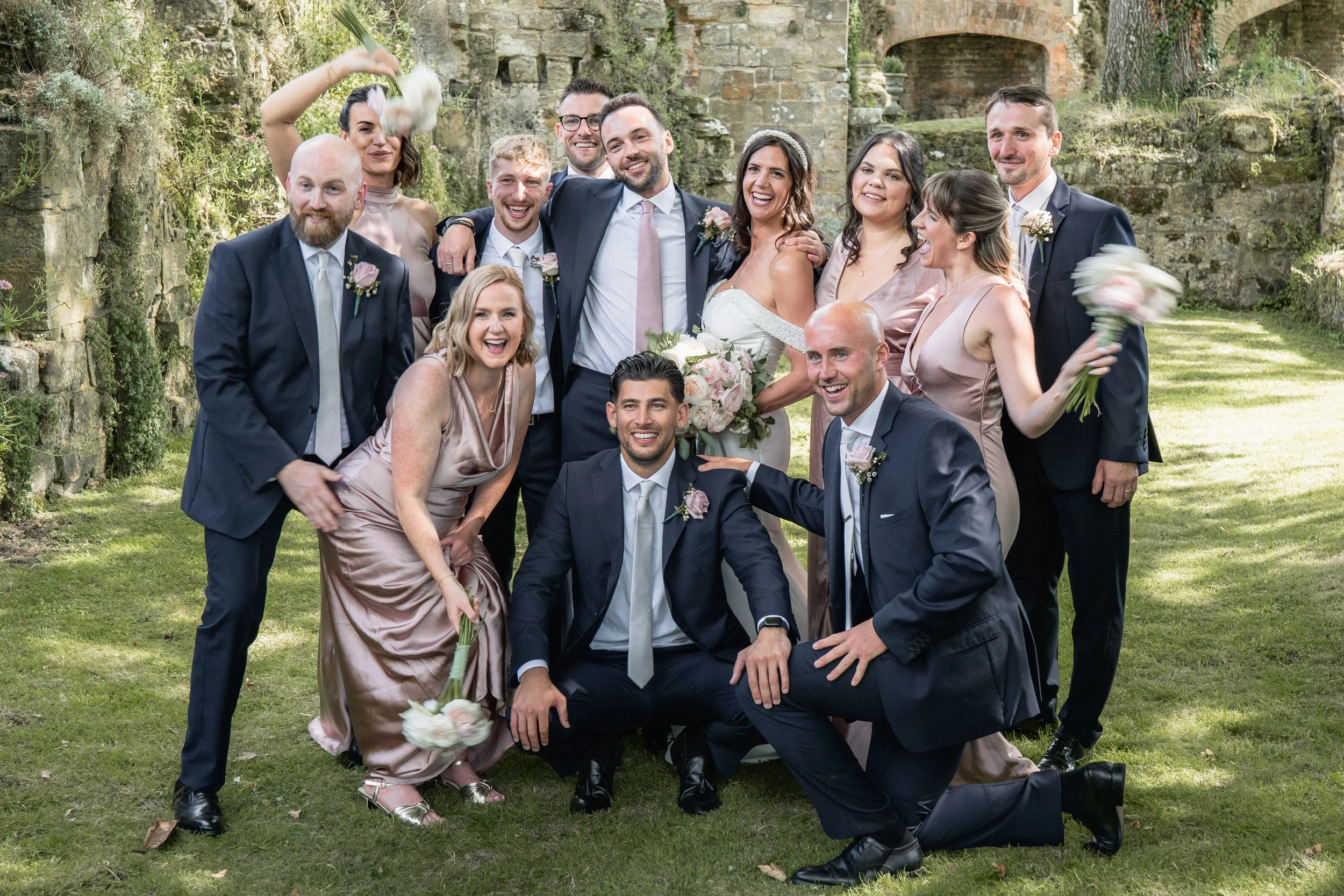 A wedding party outdoors with a group of men and women, some holding bouquets, celebrating; the bride and groom at center, smiling with friends and family around them.