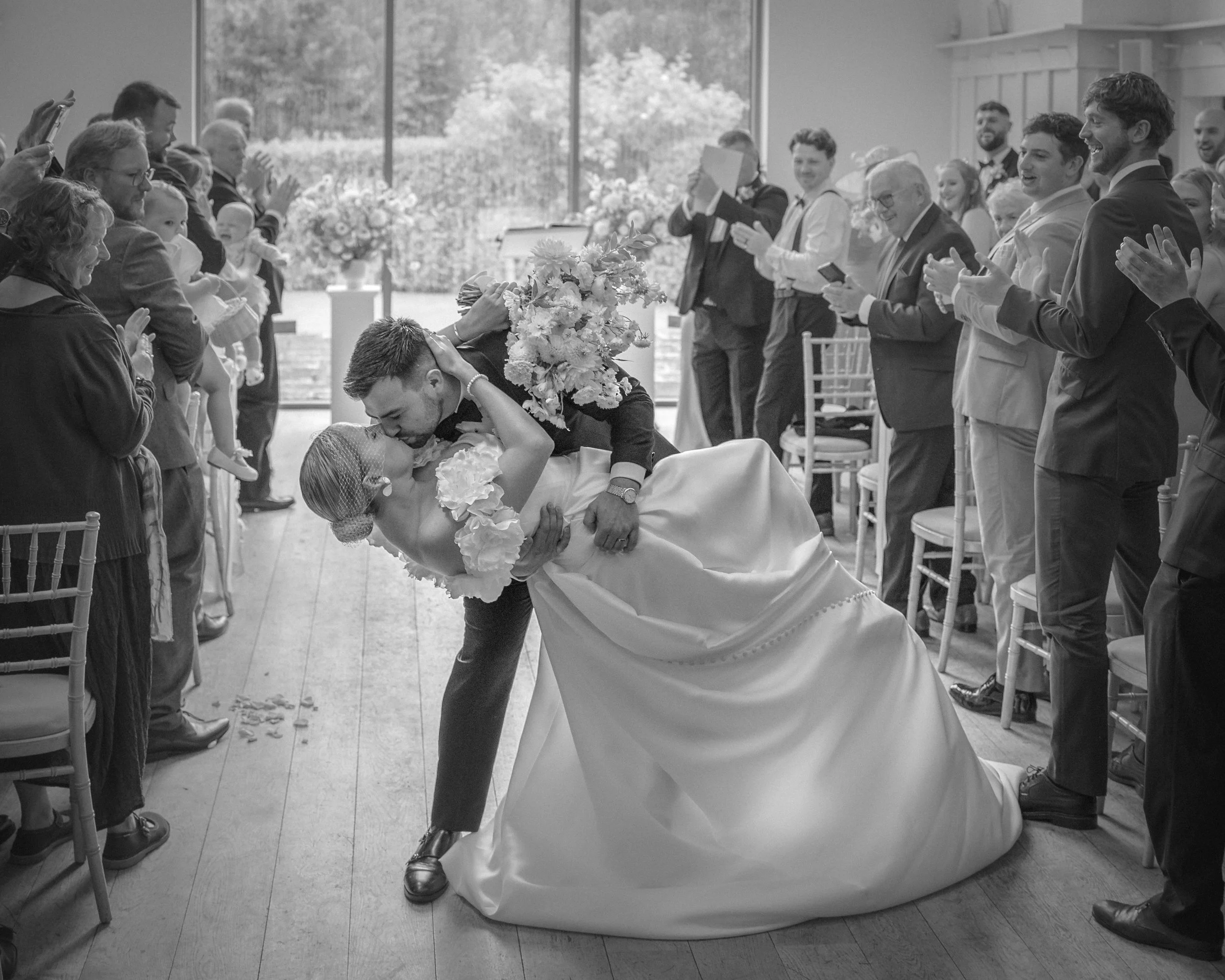 A black and white photo of a wedding reception showing a bride and groom sharing a kiss while the groom dips the bride. The bride holds a bouquet and wears a wedding gown, while the groom wears a suit. Guests are standing nearby, clapping and smiling