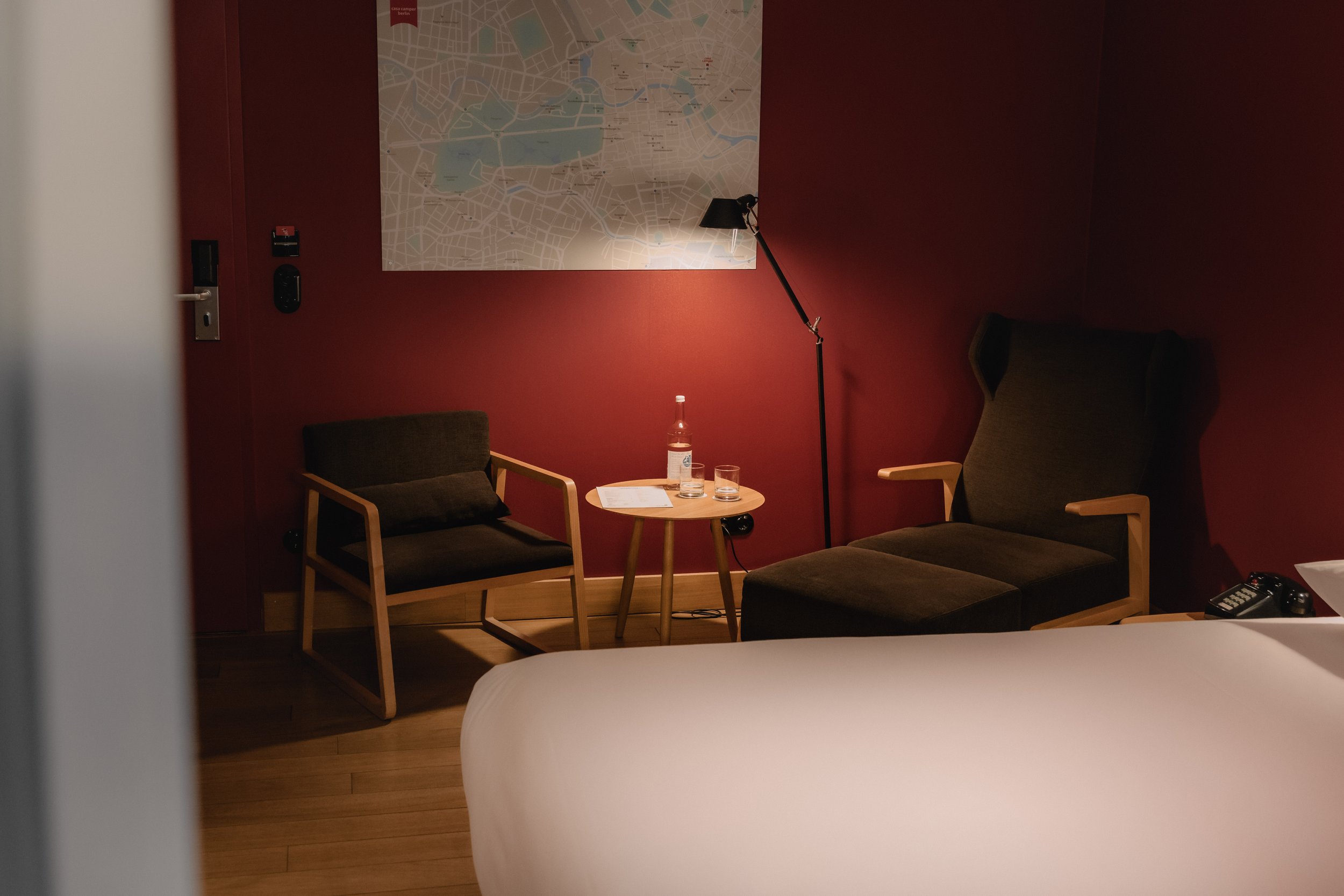 A hotel room corner with a bed in the foreground, a small round table with a water bottle, glass, and papers, two chairs, a map on the red wall, a floor lamp, and a remote control on an armrest.