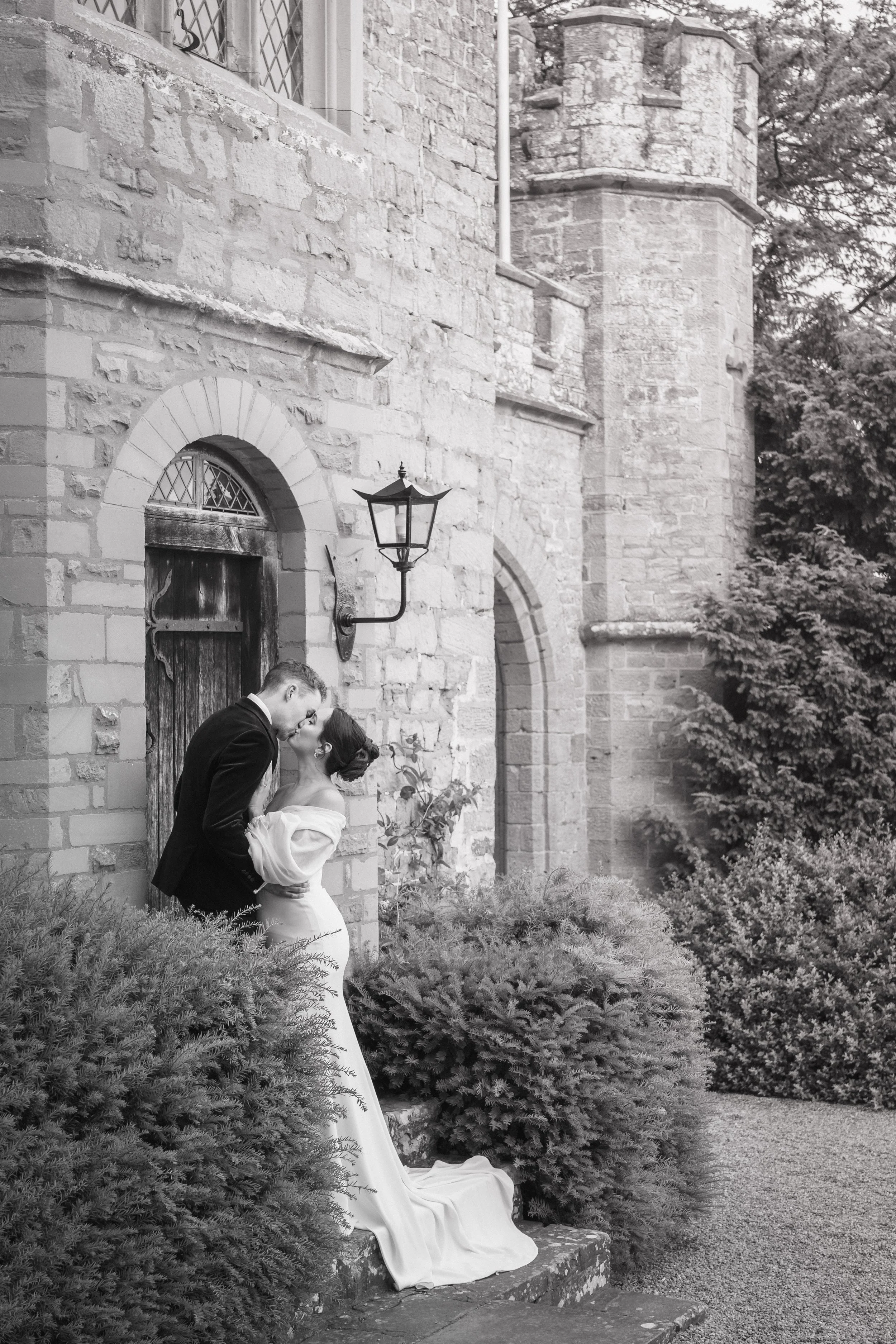 A black and white photo of a bride and groom sharing a kiss outside a stone building. The groom is in a black suit, and the bride is in a long, elegant dress. They are standing on a small stone staircase surrounded by bushes.