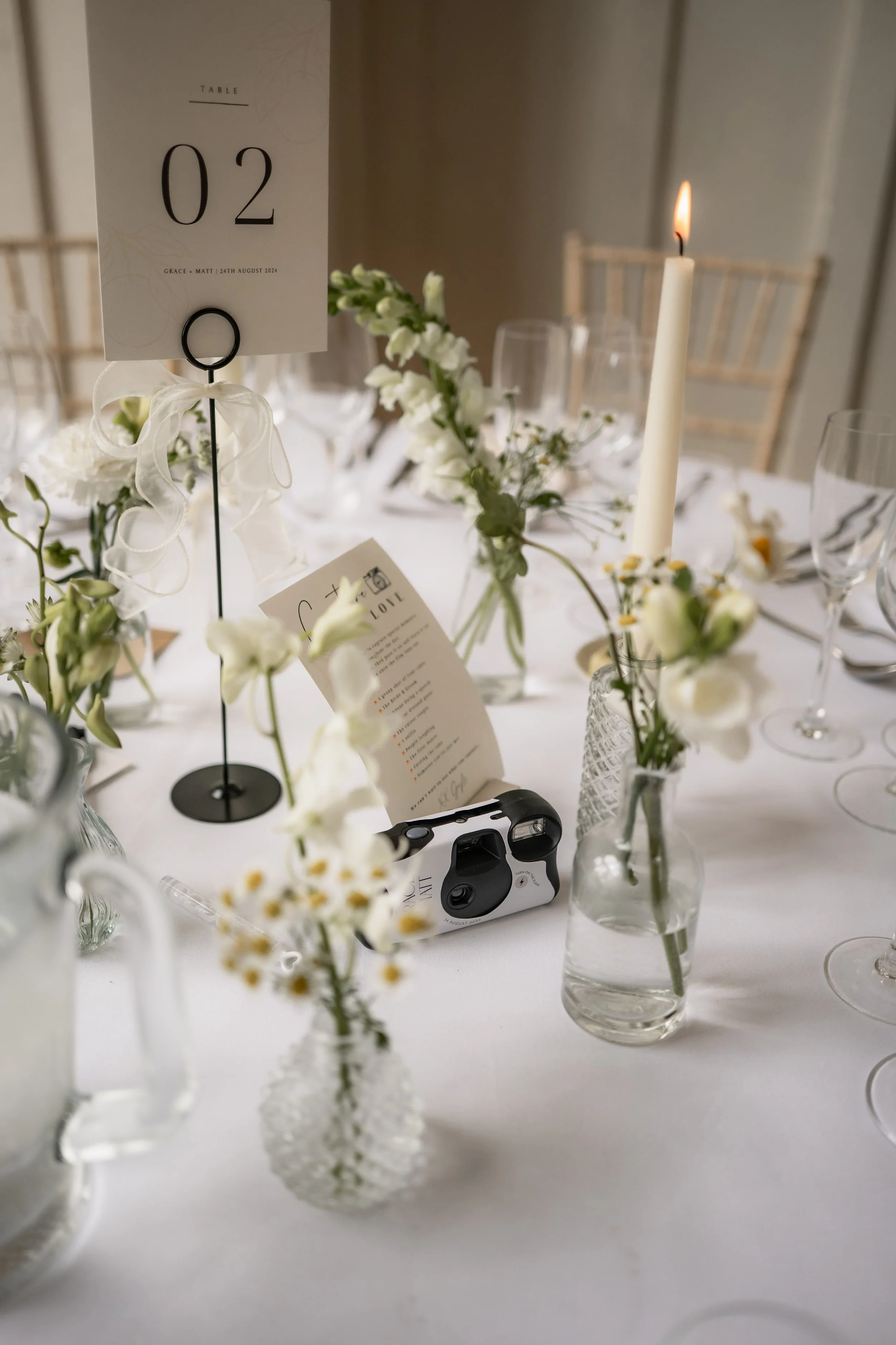 Elegant wedding reception table with white floral centerpieces, a lit candle, a table number sign, and a vintage camera, set on a white tablecloth with glassware.
