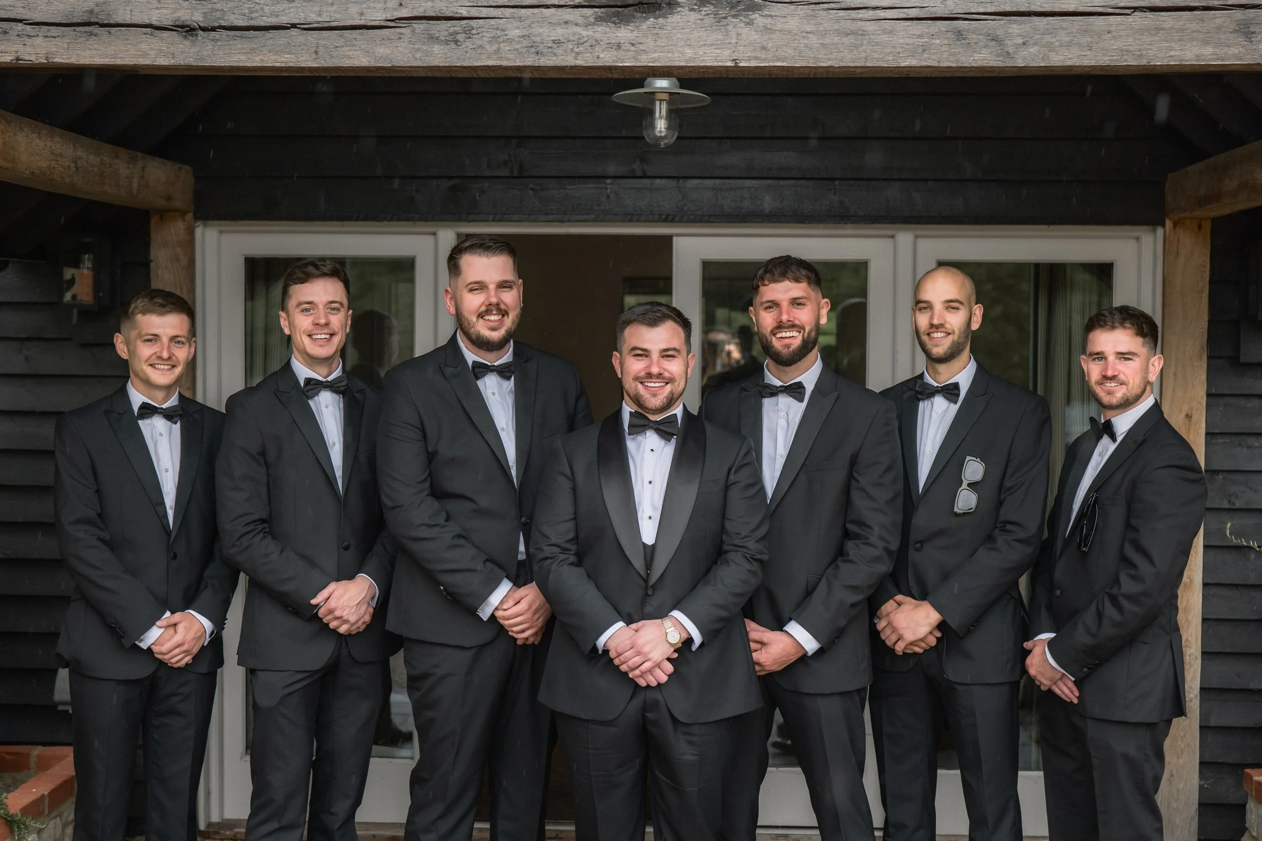 Seven men dressed in black tuxedos with white shirts and black bow ties, standing in front of a building with large glass doors, smiling at the camera.