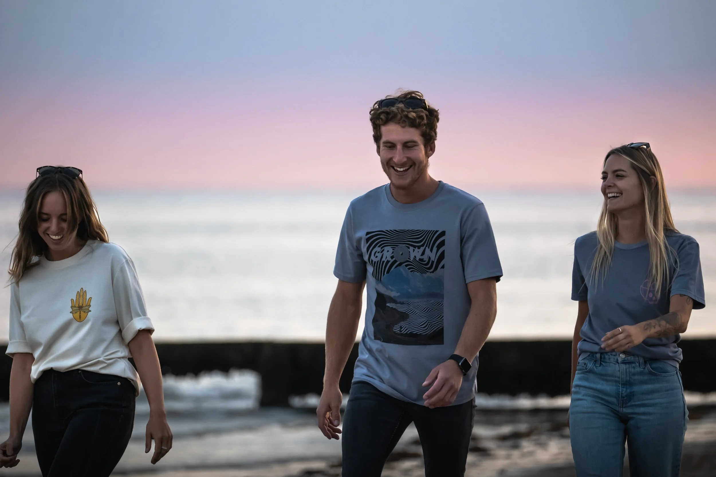 Three young adults walking and smiling on the beach during sunset, with the ocean and a pier in the background.