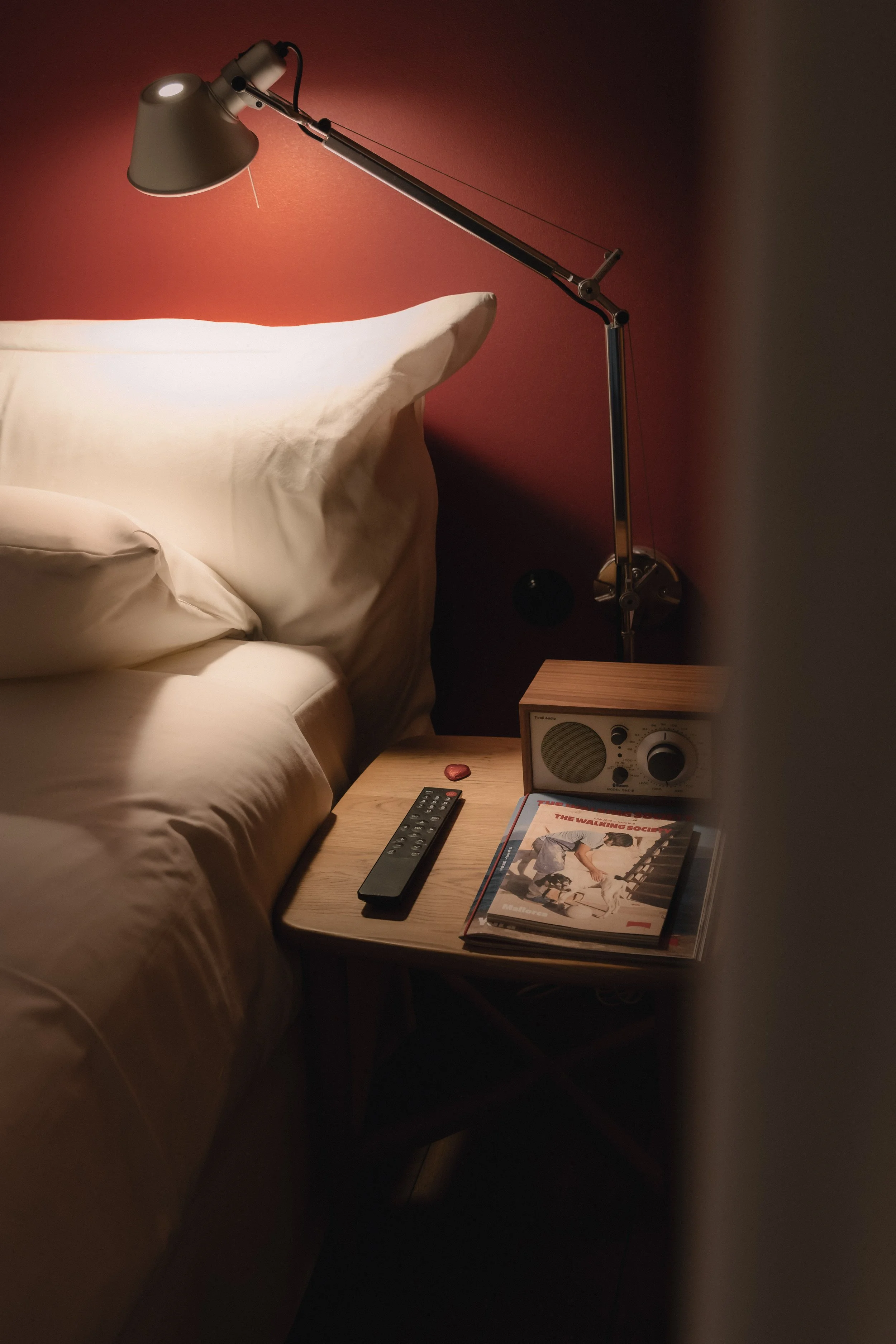 A bedside table next to a bed with beige sheets and pillows, with a modern adjustable desk lamp, a remote control, a vintage-style radio, and a vinyl record album on top of the table, with a red wall in the background.