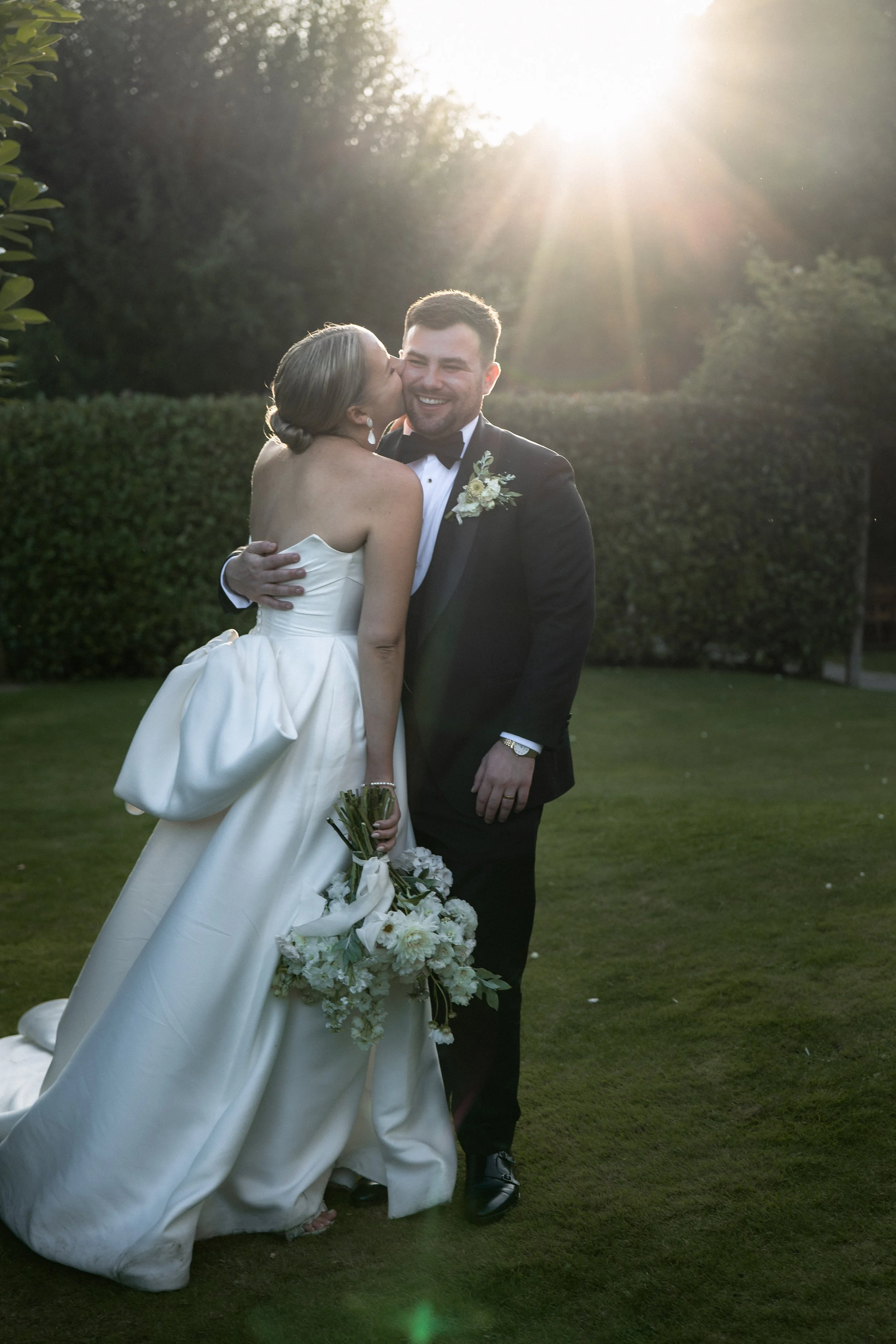 A bride and groom sharing a kiss outdoors at sunset, with the bride holding a bouquet of white flowers and the groom smiling, surrounded by greenery.