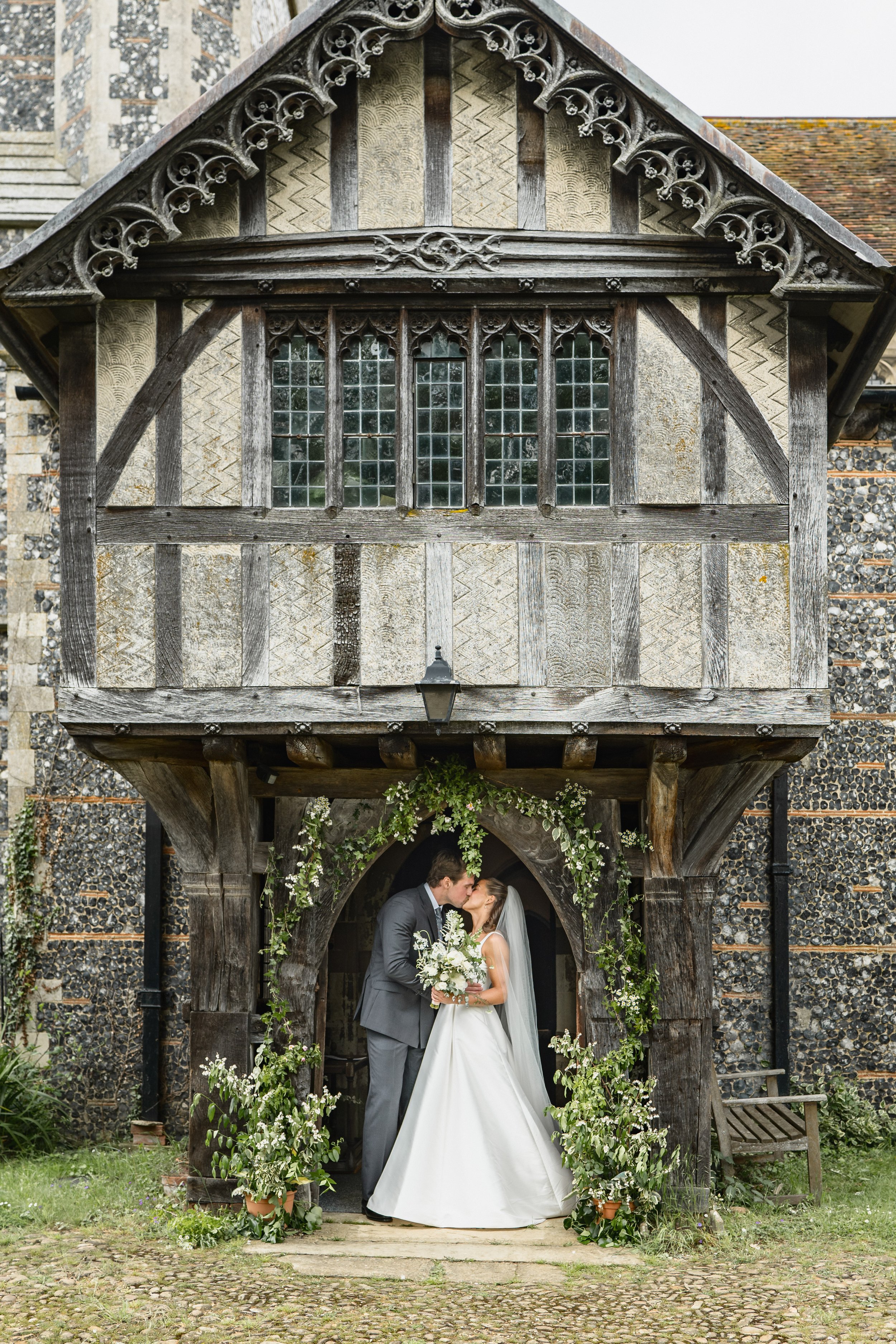 A bride and groom kissing beneath an arch decorated with white flowers in front of a rustic wood and stone building.