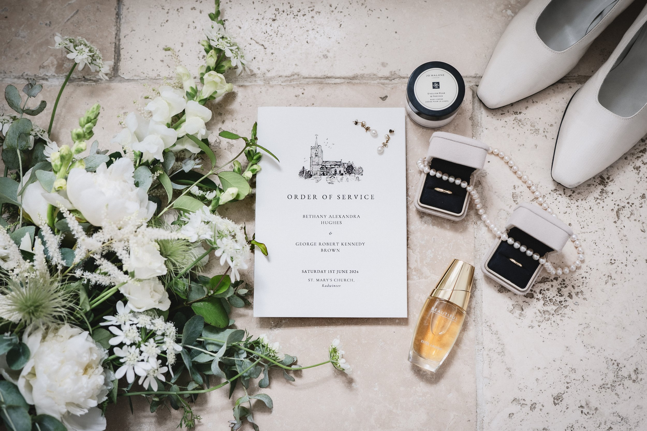 Wedding order of service invitation surrounded by white flowers, pearl jewelry, a bottle of perfume, pairs of white shoes, and jewelry boxes.