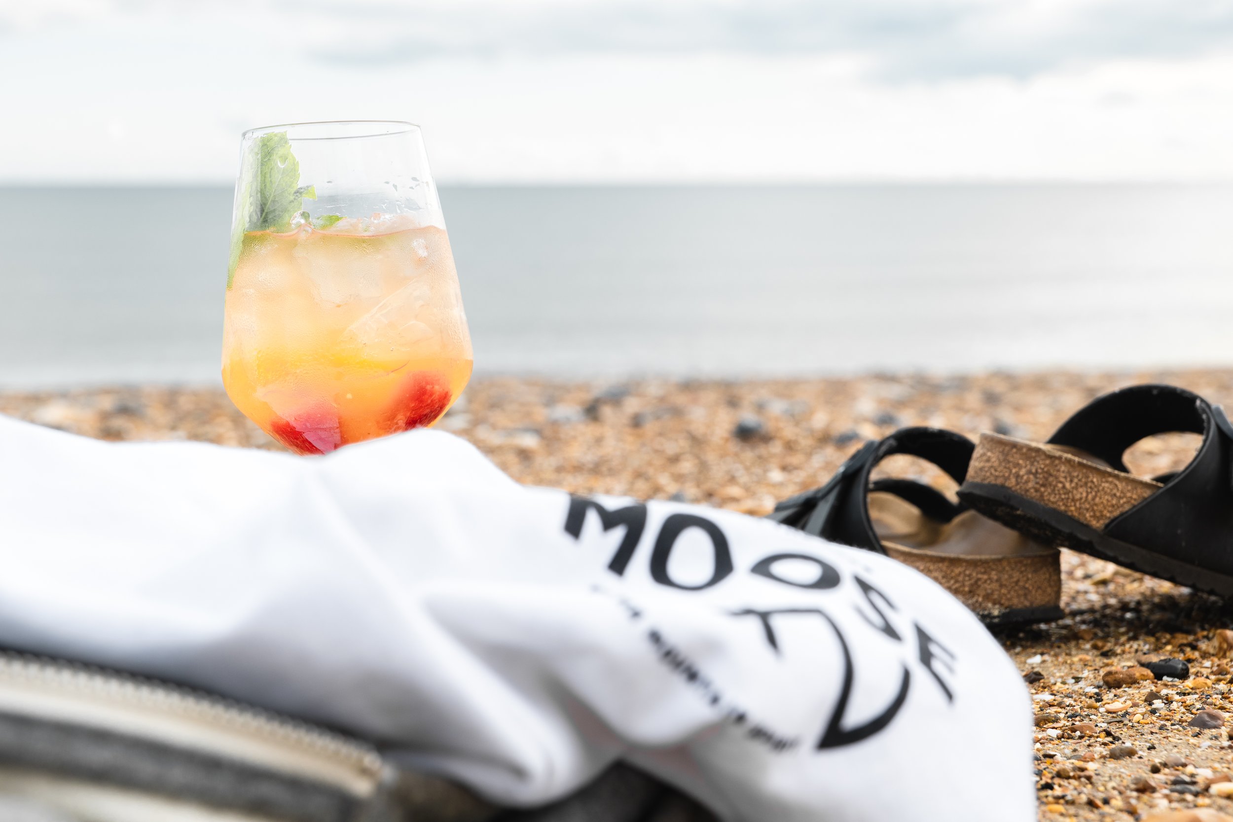 A tropical cocktail with fruit and ice in a glass on a beach, with black sandals and a white cloth with text, and a calm ocean in the background.