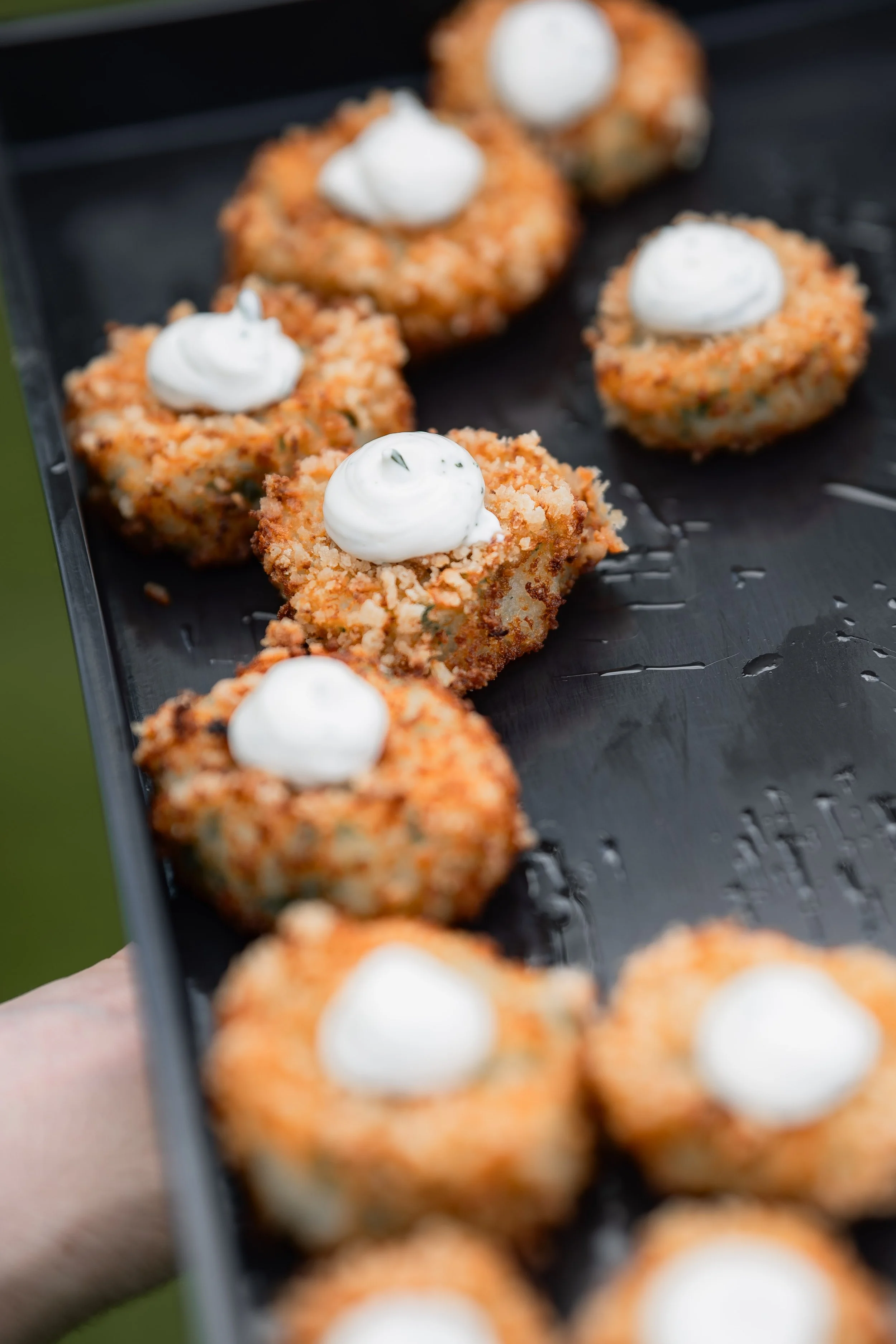 Close-up of breaded and baked or fried bite-sized appetizers topped with dollops of white creamy sauce, arranged on a black serving tray.
