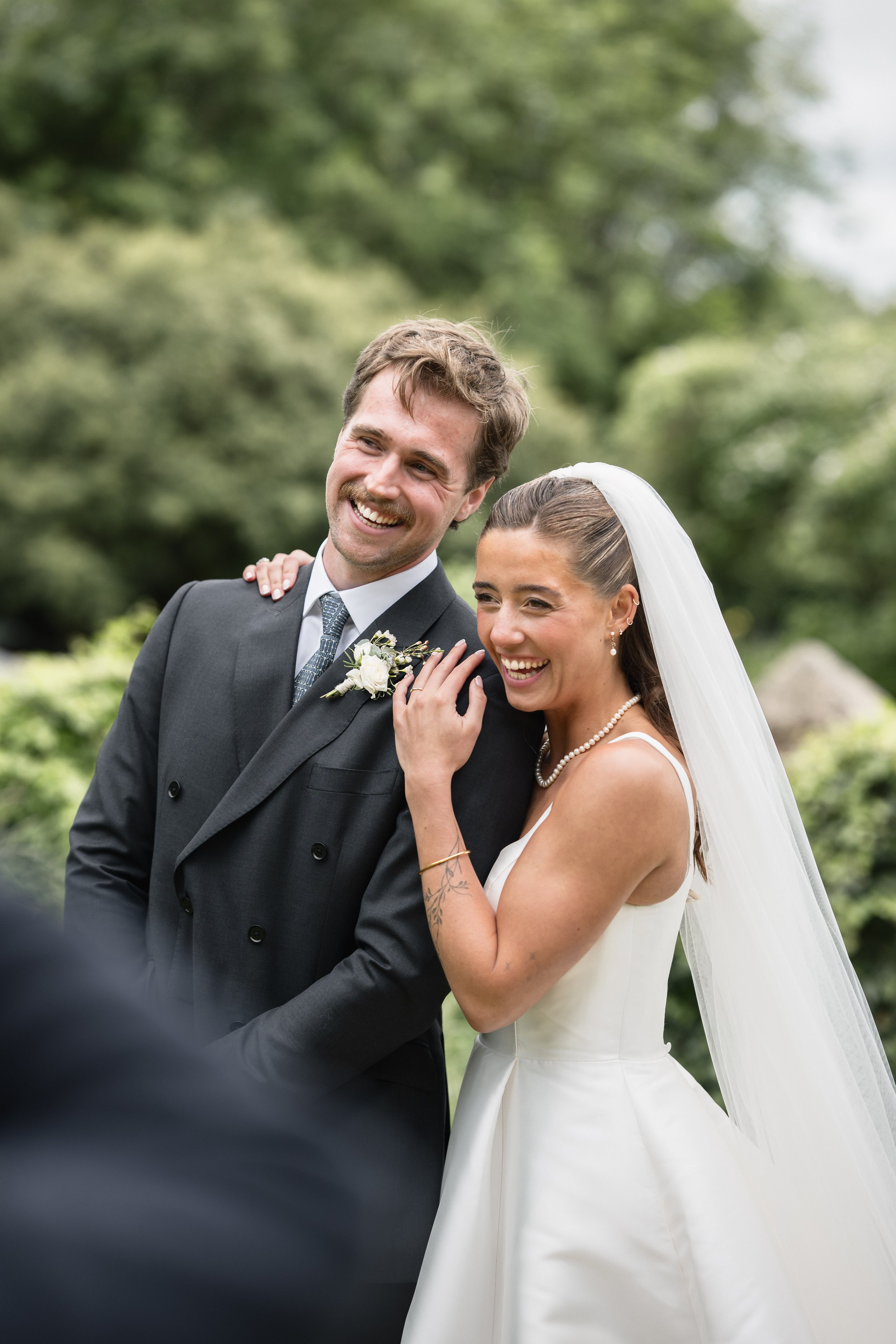 Smiling bride and groom at outdoor wedding, with the bride wearing a white dress and pearl necklace, and the groom in a dark suit with a white boutonniere, standing in a green, natural setting.