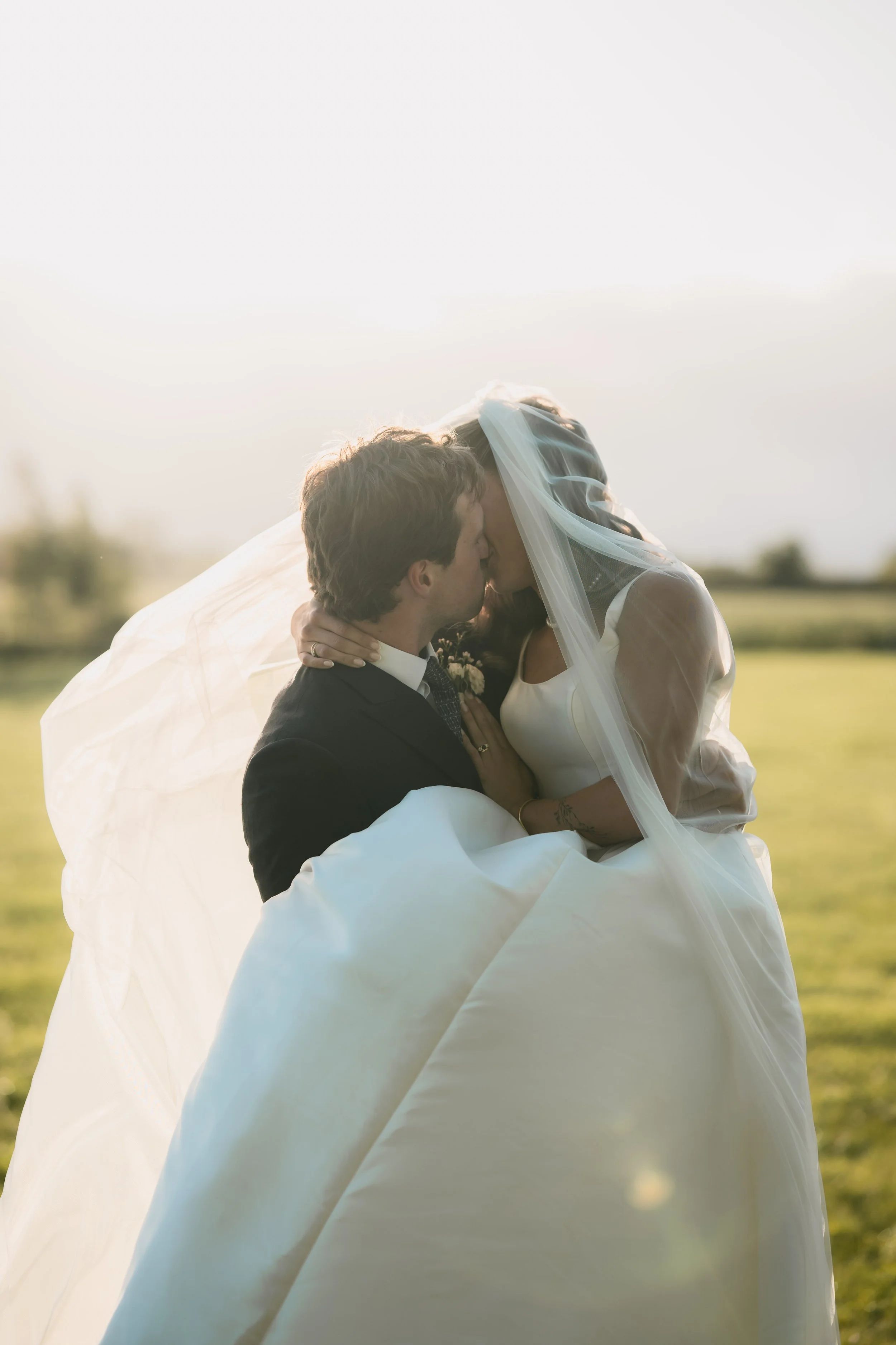 A bride and groom sharing a kiss outdoors, with the bride in a white wedding dress and veil, and the groom in a black suit, during sunset.