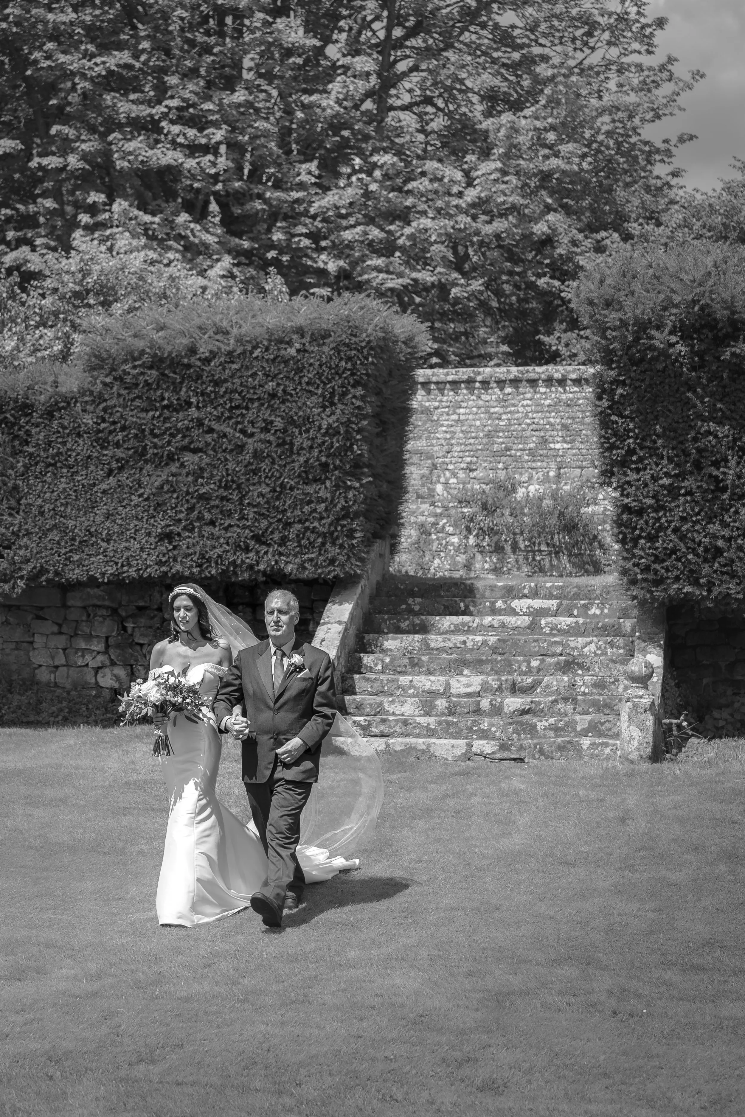Black and white photo of a bride in a wedding dress with a veil and bouquet, walking arm-in-arm with a groom in a suit, outdoors near stone stairs and trimmed hedges.
