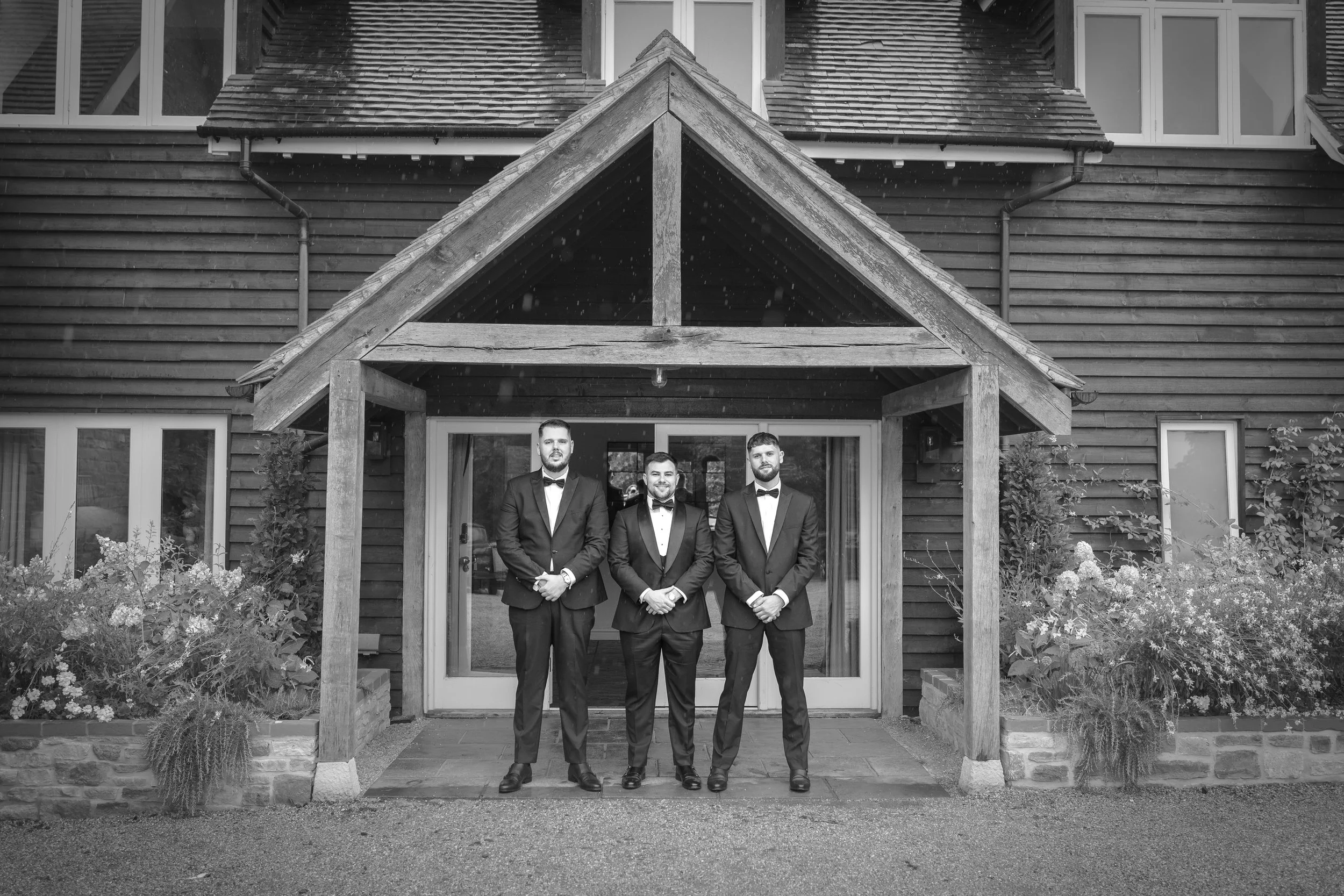 Three men in tuxedos and bow ties standing outside of a house with wooden siding and a small porch, in black and white.