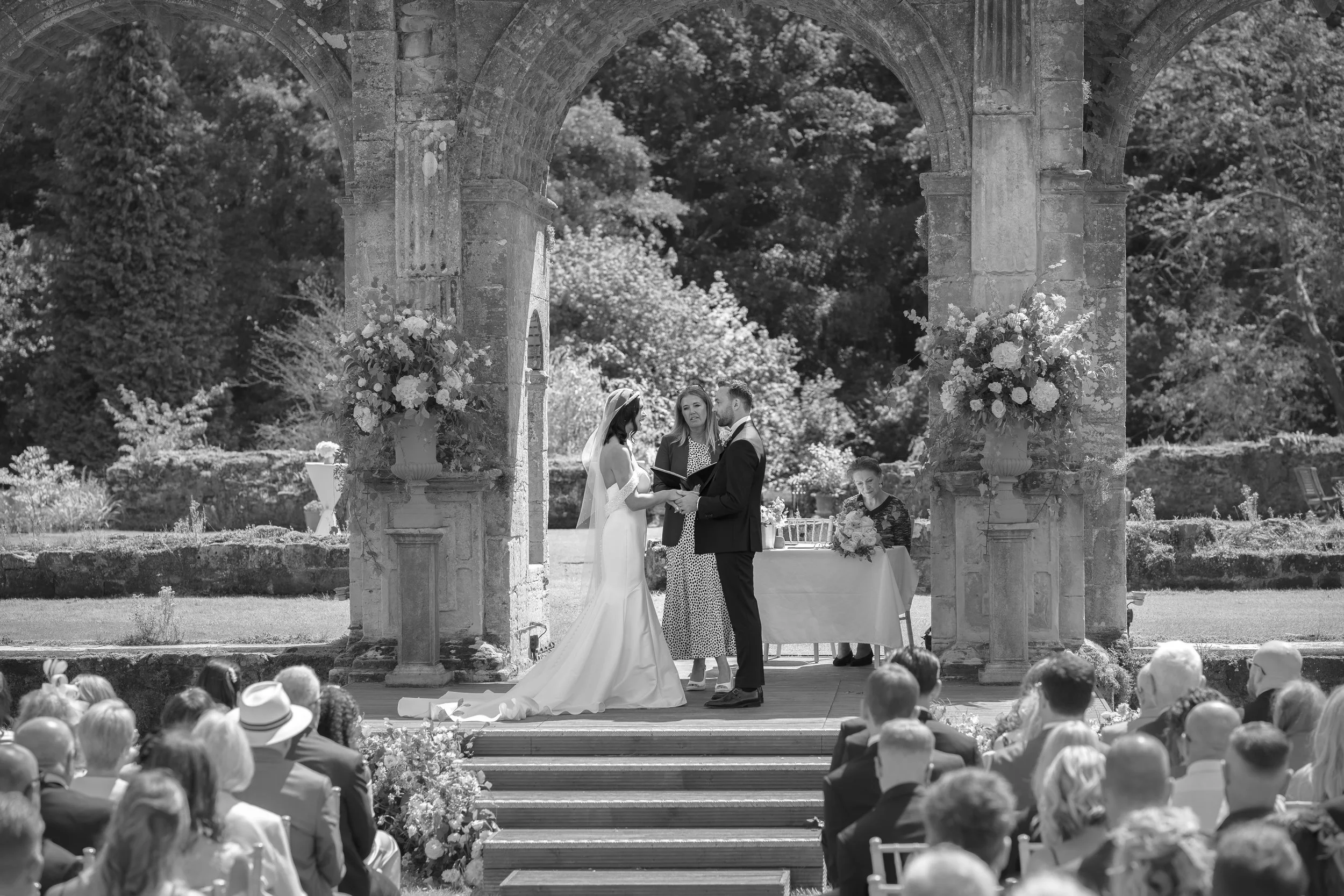 A black and white photograph of a wedding ceremony taking place outdoors under a stone archway with guests seated in front. The bride and groom stand facing each other, holding hands, with a woman officiating behind them. There are floral arrangement