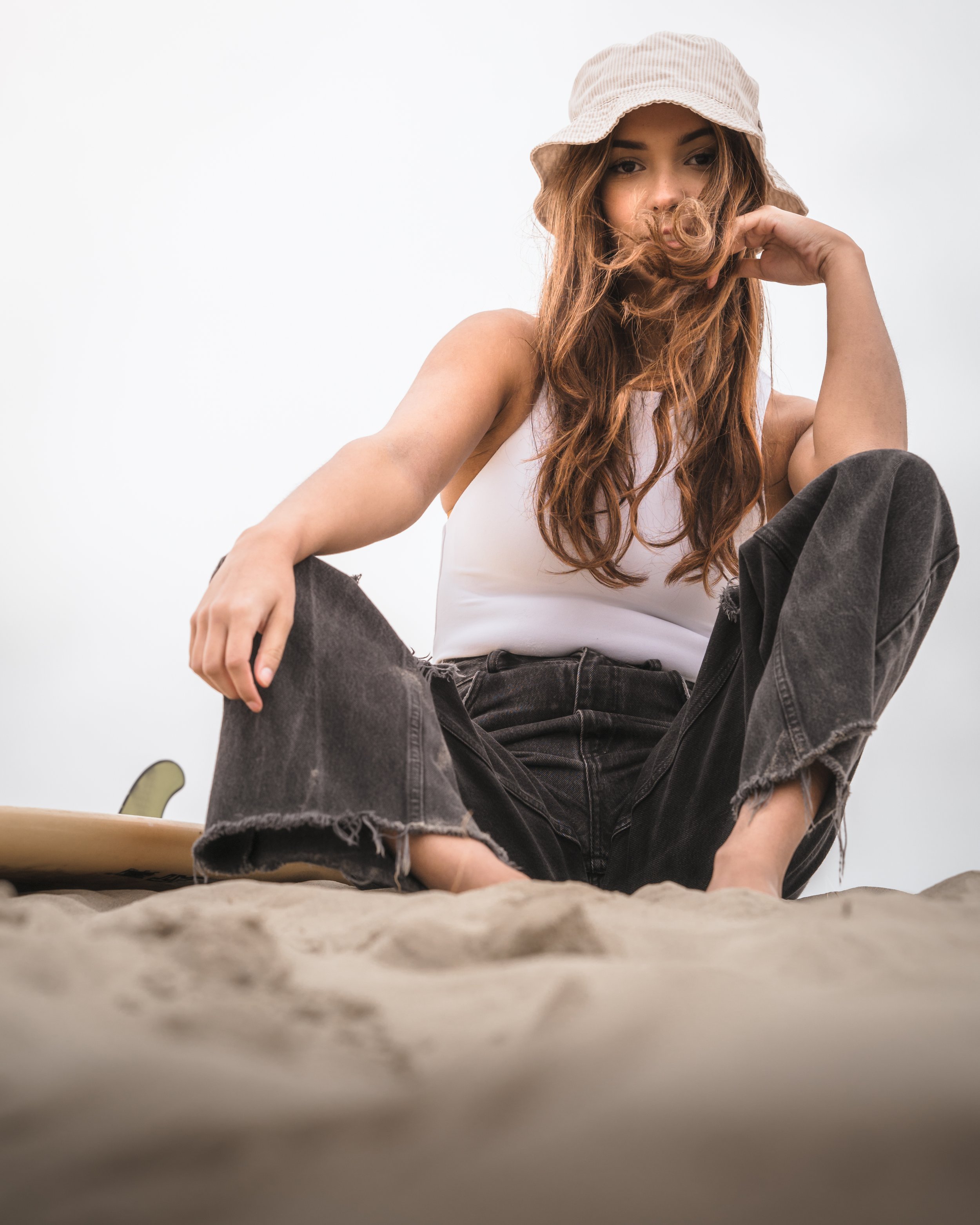 Young woman sitting on sandy beach, wearing a beige bucket hat, white tank top, and black ripped jeans, with wavy brown hair partially covering her face.
