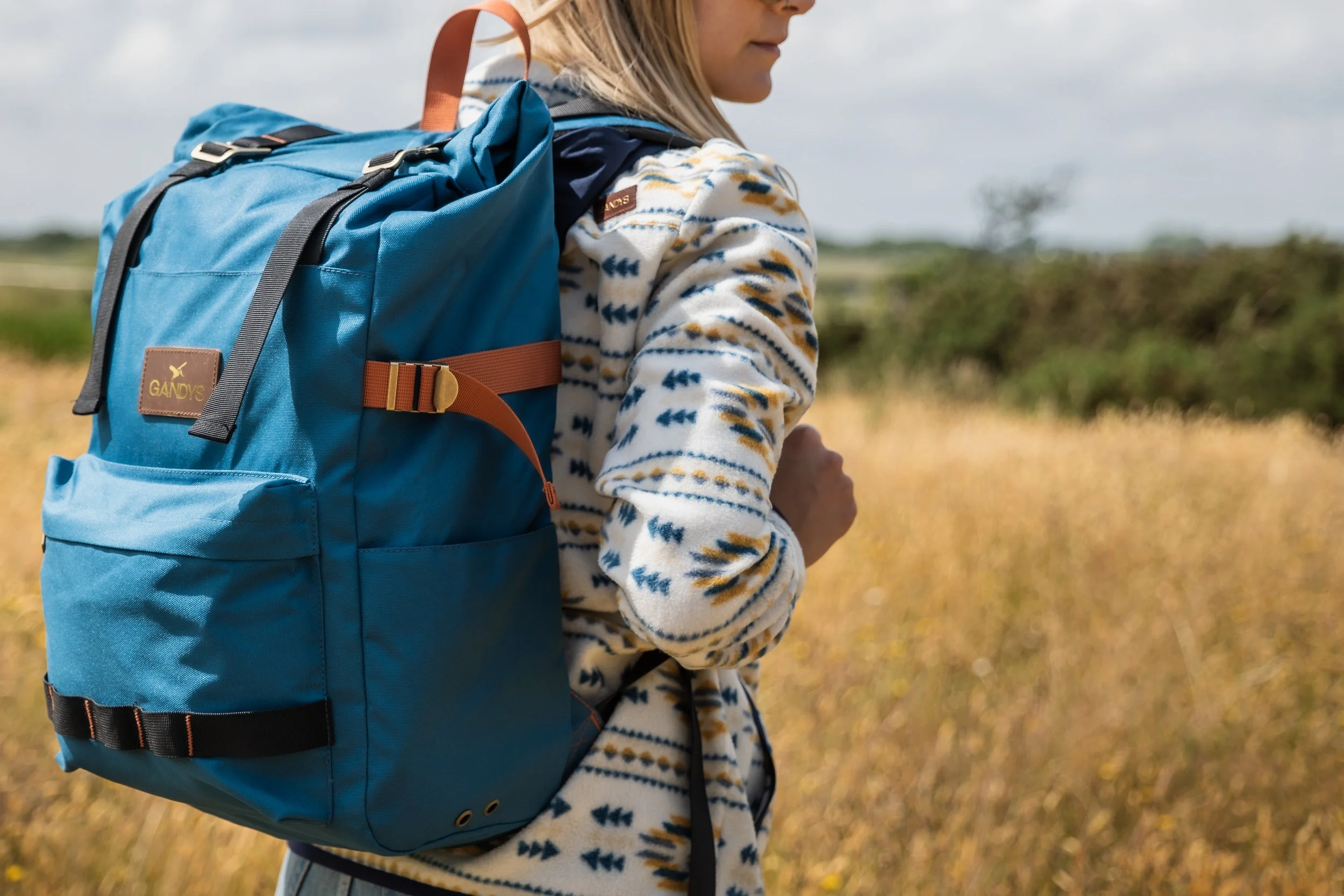 A woman with a large blue backpack walking through a field of tall grass with trees in the background.