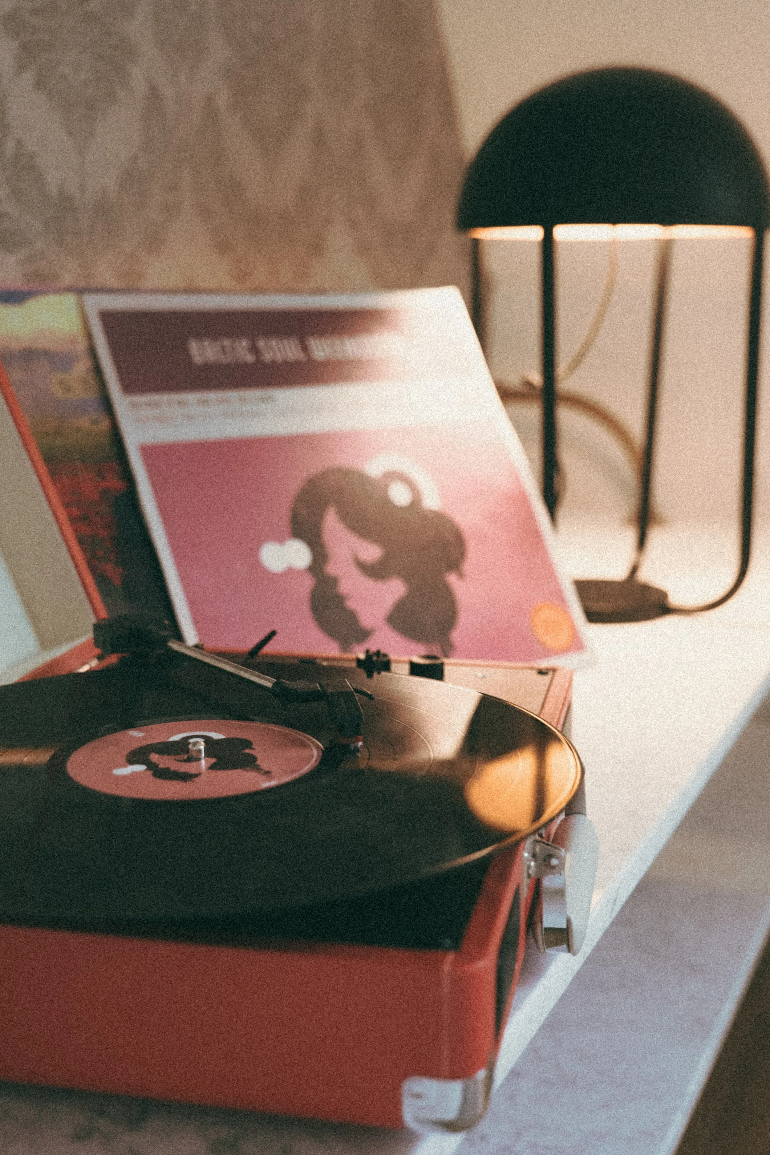 A vintage red record player playing a vinyl with a pink album cover visible in the background, and a black desk lamp with a semi-rounded shade on a white surface.