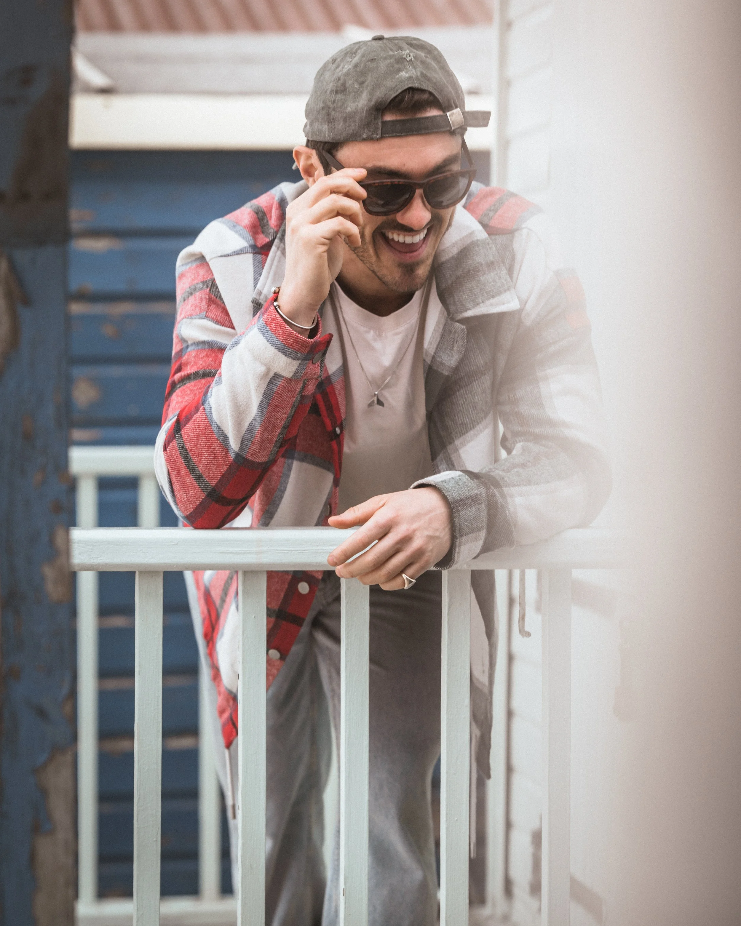 Young man wearing a grey cap, sunglasses, a red plaid jacket, and a white t-shirt leaning on a white porch railing, smiling and talking on a mobile phone.